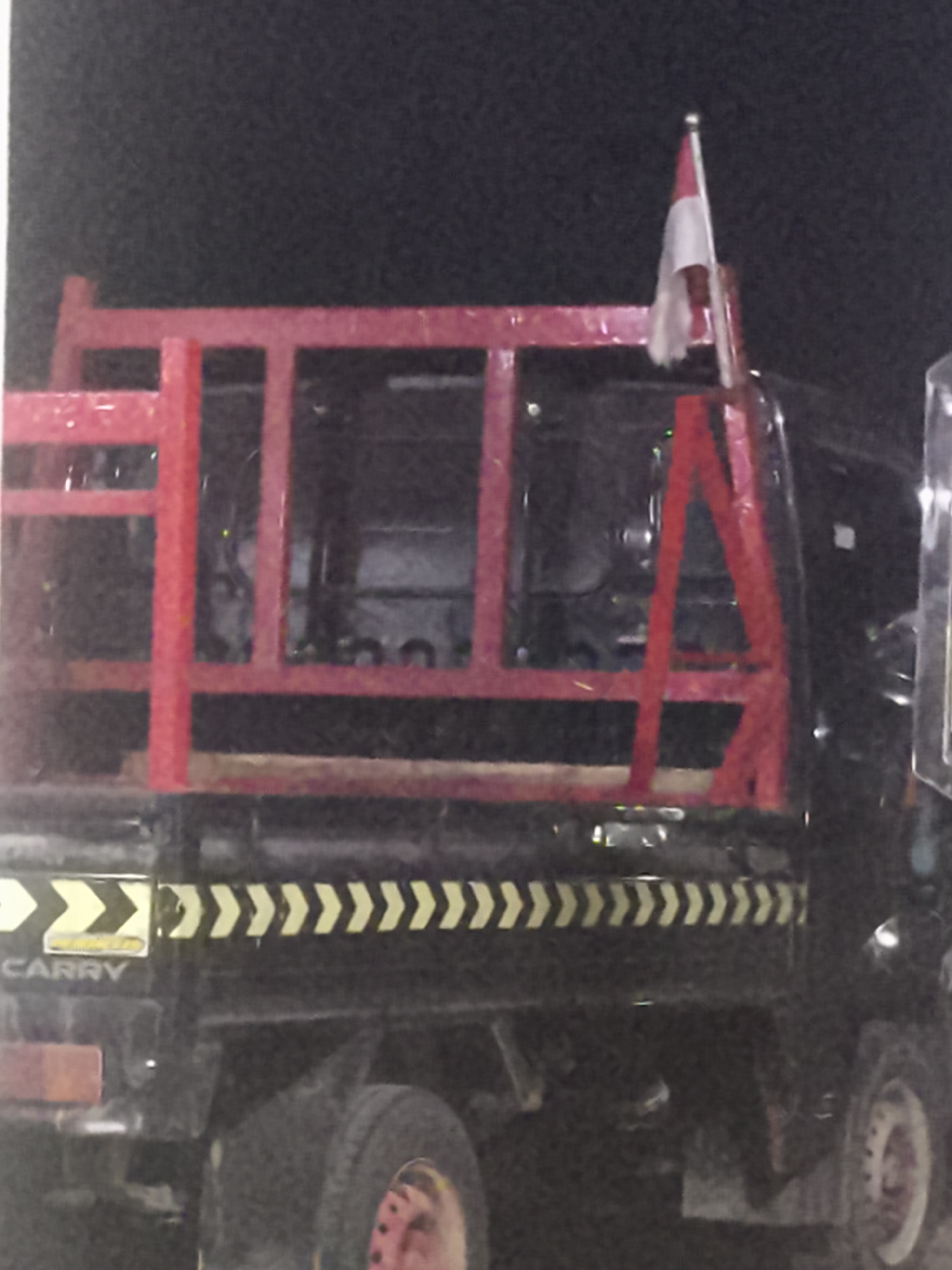 An unidentified vehicle, likely a truck, is parked in an outdoor environment at night. The vehicle has a red metal frame structure loaded onto its bed, which appears to be made of dark material. A small flag, white and red, is attached to a pole on the top right of the red frame. The side of the vehicle has a strip of alternating yellow and black chevron markings. Text that reads "CARRY" is visible on the side of the truck, towards the rear. The wheels of the truck are visible. The scene is dimly lit, with the background appearing dark and indistinct, suggesting it is nighttime. The image lacks any discernible architectural features or natural landmarks that would help pinpoint a specific location within Samarinda, Indonesia, or any other city.