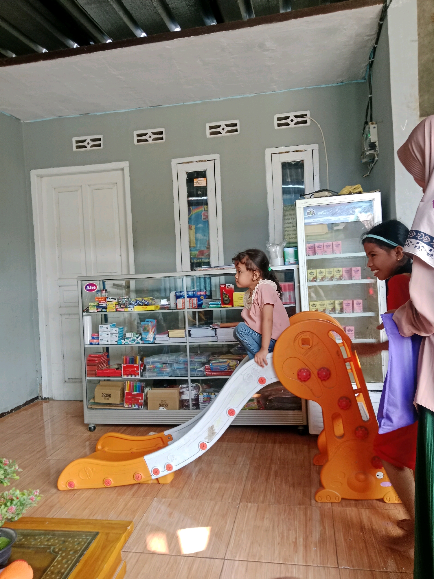 In Ciranjang, Indonesia, within a room featuring a grey painted wall, a white door, and a corrugated metal ceiling, a young girl with dark hair tied in a ponytail sits on the top of a small, orange and white plastic slide. She is wearing a pink t-shirt and blue jeans and is looking to her left. To her right, another young girl with dark hair and a blue headband, wearing an orange t-shirt and a pink hijab, smiles while holding a purple cloth. In the background, there is a glass-fronted display cabinet filled with various items, including stationery and packaged goods. A second, taller display cabinet on the right holds rows of small boxes. The floor is covered in light brown tiled linoleum.