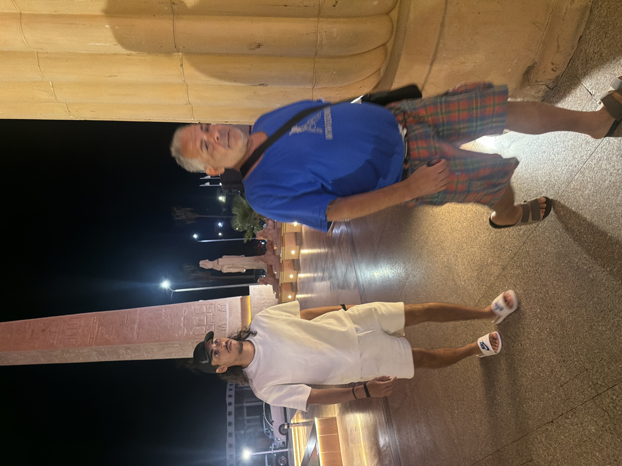 Two men are walking on a paved pathway at night, with large illuminated structures in the background. The man on the left is wearing a white t-shirt, white shorts, and white slide sandals. He has dark hair and is wearing a black baseball cap. The man on the right is wearing a blue t-shirt, plaid shorts, and dark slide sandals. He has grey hair and is carrying a black bag over his shoulder. Behind the men, there is a tall, illuminated obelisk with hieroglyphics. Further back, there are more illuminated structures, palm trees, and streetlights. The scene appears to be in a public area, possibly a tourist attraction or a promenade in Sharm El Sheikh, Egypt, given the presence of Egyptian-style monuments.