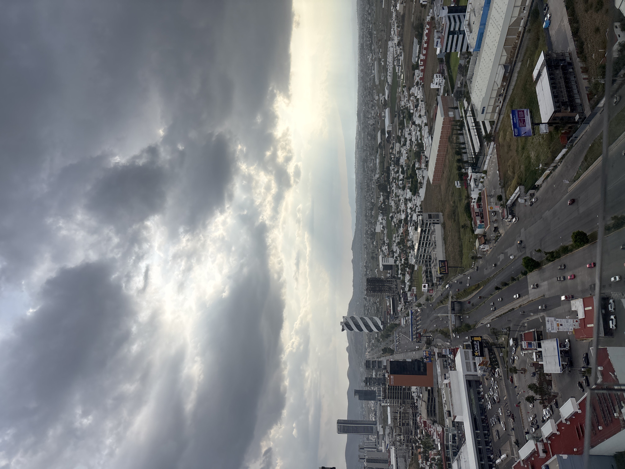 The image shows an aerial view of San Bernardino Tlaxcalancingo, Mexico, under a cloudy sky. The cityscape is visible, featuring a mix of residential and commercial buildings, roads with vehicular traffic, and open green spaces. In the distance, there are mountains partially obscured by atmospheric haze. The overall scene suggests a densely populated urban area with a diverse architectural landscape.