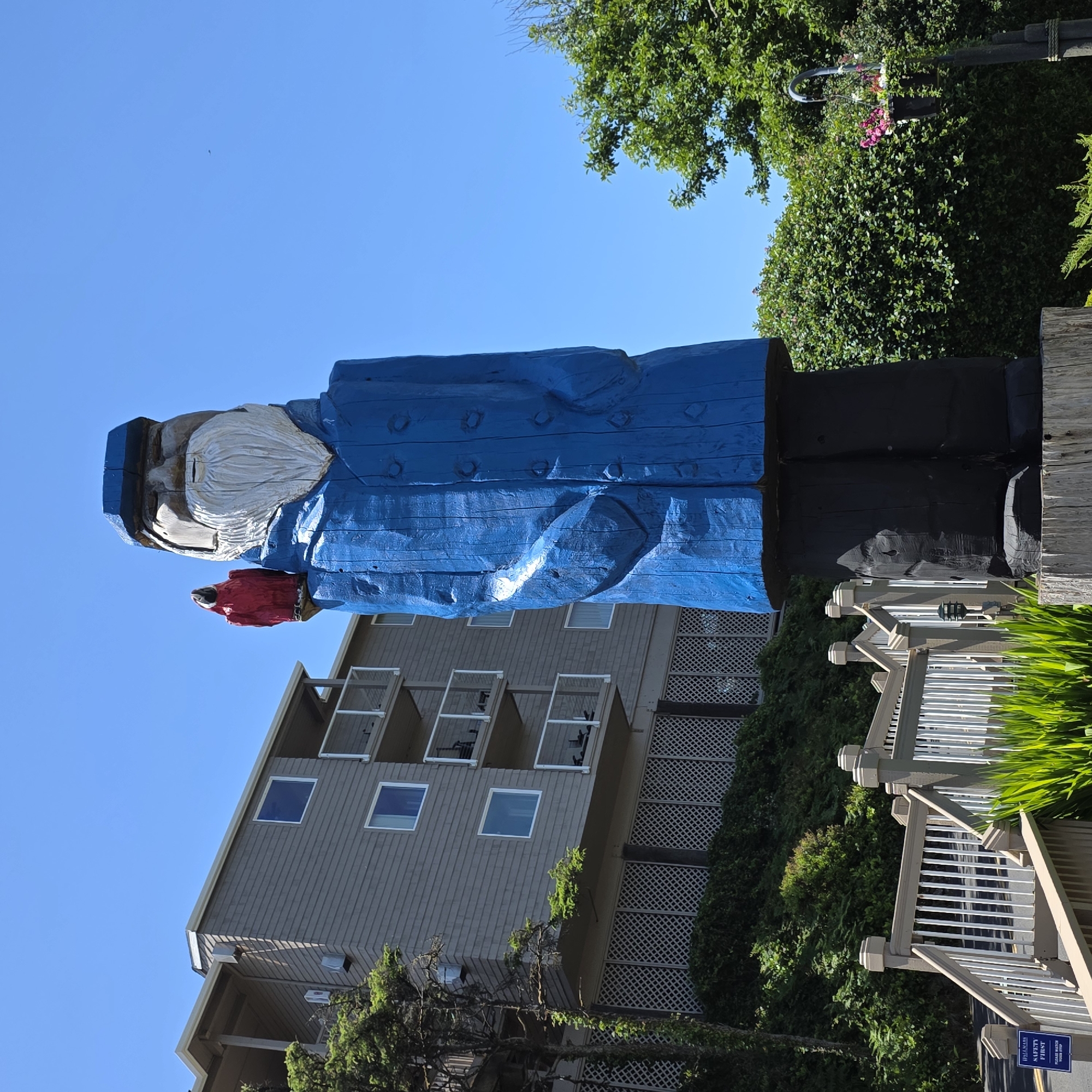 This image captures a towering wooden sculpture of a fisherman in Cannon Beach, Oregon, USA. The sculpture, painted in vibrant blue and black, depicts a man with a white beard, wearing a cap and a coat with visible buttons. A small red bird is perched on his shoulder. In the background, a multi-story building with gray siding and balconies is visible, nestled amongst lush green trees and foliage. A white wooden railing and stairs lead up towards the building, with some greenery in the foreground. The sky above is a clear, bright blue.