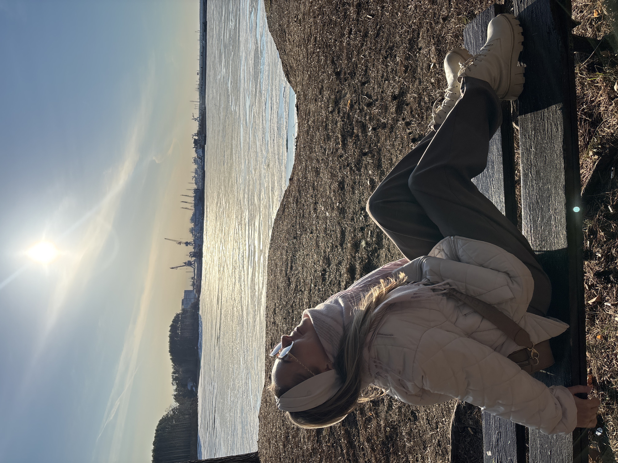 A photograph shows a young woman reclining on a bench, her feet pointing towards the top right of the frame. She is wearing a white quilted jacket, brown trousers, and off-white platform boots. Her hair is blonde, and she has sunglasses perched on her face. A bag with a brown strap rests on her torso. The background reveals a body of water, likely the Kama River, with a cityscape visible in the distance, suggesting the city of Chaikovsky, Russia. The sky is pale blue, with the sun casting a bright glare. The bench sits on a grassy or dirt patch.