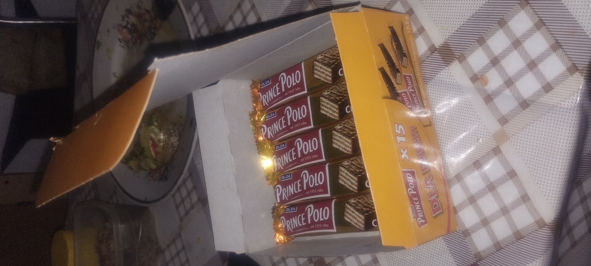 A box of Prince Polo wafers is shown in an indoor setting. The box is yellow and red, with the Prince Polo logo prominently displayed. Inside the box, there are several individually wrapped wafers, each with a chocolate filling and wafer exterior. Some of the wafers are visible from the top, showing the golden foil wrapping. The box appears to be full. The background is blurred, but suggests a table covered with a checkered tablecloth. There are also some other food items visible in the background, including a plate with what looks like a salad or appetizer. The lighting is dim, with a bright reflection on the foil of the wafers.