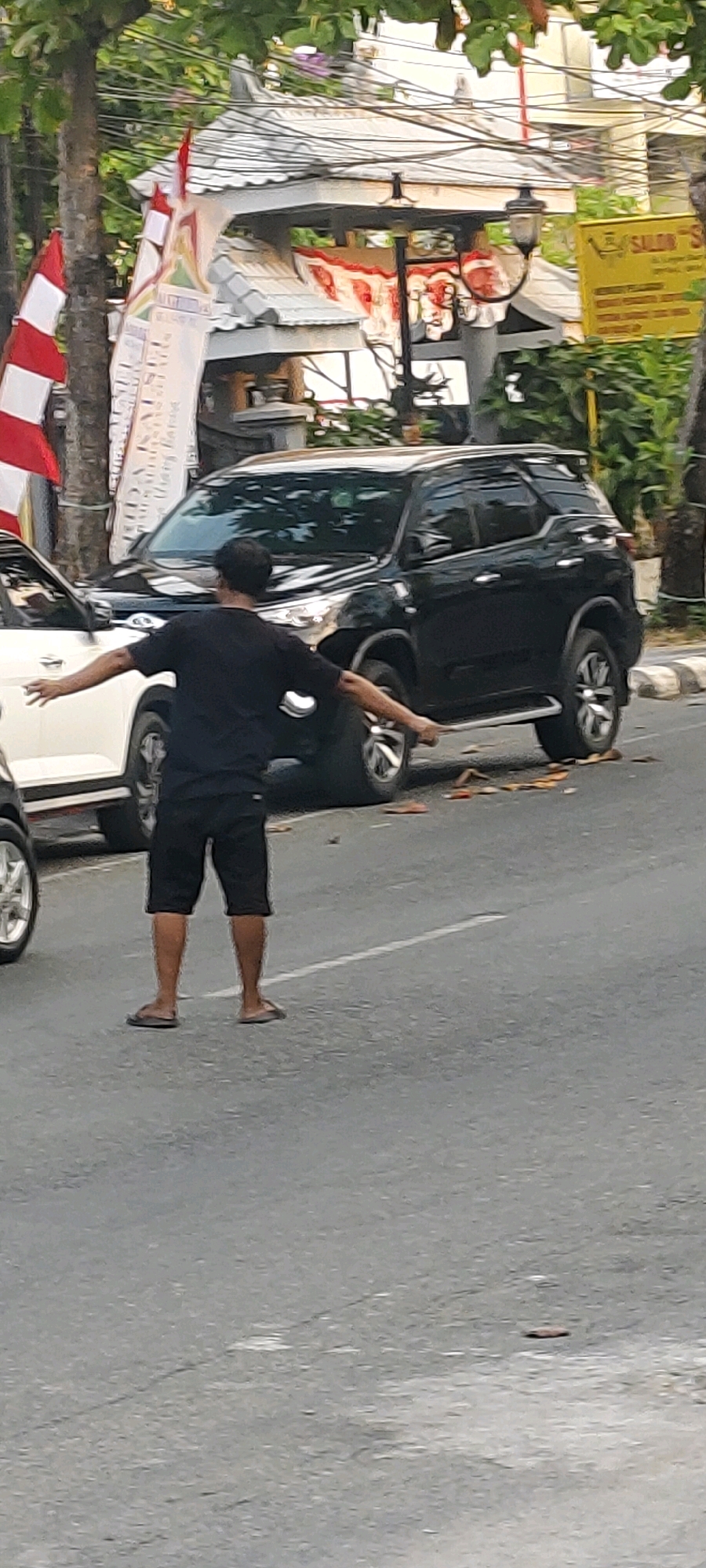 A man in a black t-shirt and black shorts stands in the middle of a paved road, with his arms outstretched. He is wearing flip-flops. Two cars are parked on the road beside him; a white car on the left and a black SUV on the right. In the background, there are trees, a building with a white tiled roof and red and white flags, and a yellow sign that reads "SALON'S." The scene appears to be in a city, possibly Banjarmasin, Indonesia, given the presence of the Indonesian flag colors on the flags. The road has a white line painted down the center. Fallen leaves are scattered on the road around the black SUV.