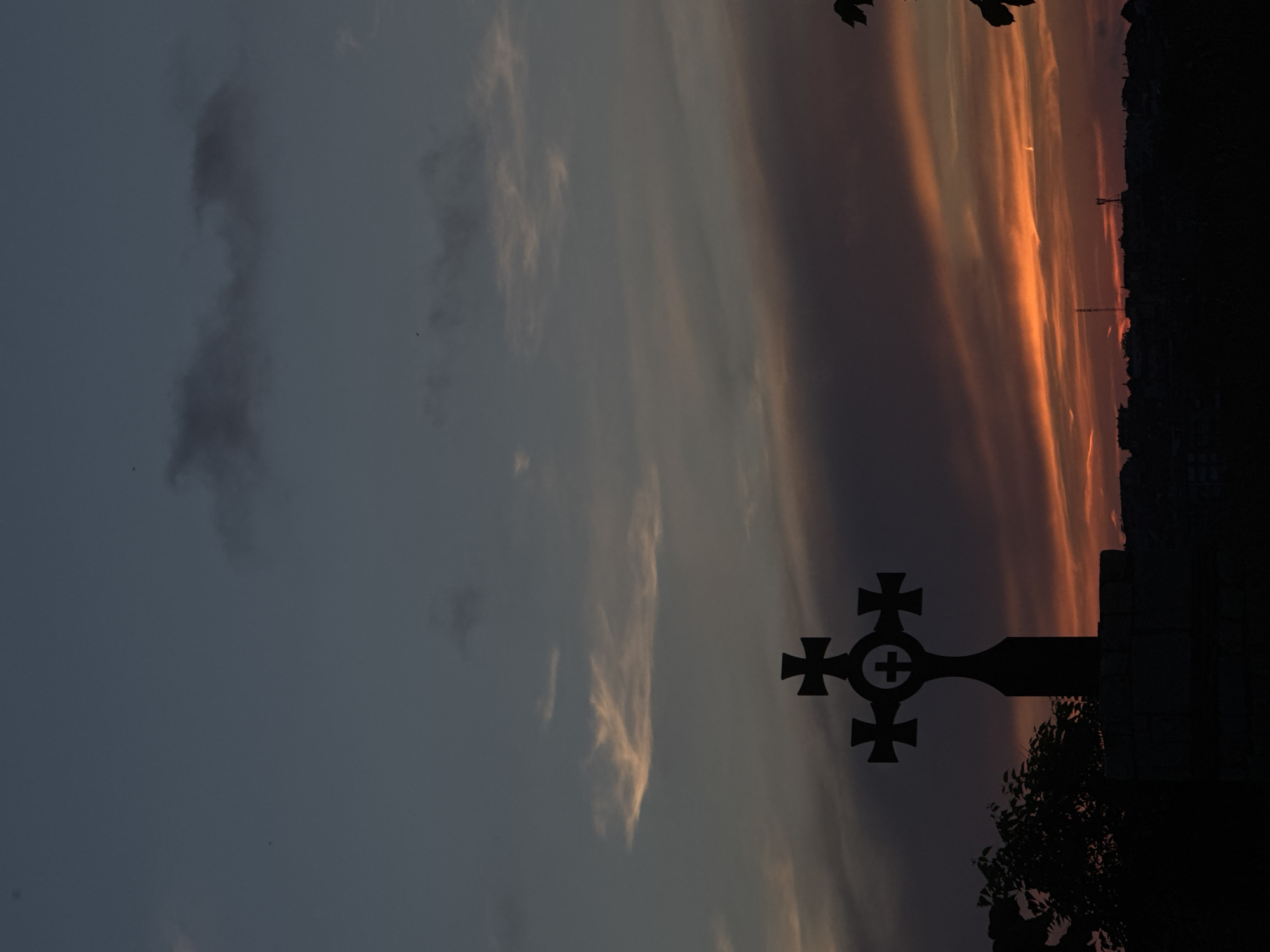 A silhouette of an elaborately designed cross stands against a dramatic sunset sky, casting a dark shadow over a low-lying town or village in the distance. The sky is a canvas of deep blues and purples, streaked with vibrant orange and yellow clouds, suggesting the sun has just dipped below the horizon. The cross, situated in the foreground, is intricately carved, featuring smaller crosses at the ends of its main beams and a circular motif in the center with a further cross etched within. To the left of the cross, the dark silhouette of foliage is visible. The town in the background is a jagged line of buildings, with indistinct shapes of structures and possibly communication towers piercing the twilight. The overall atmosphere is somber and evocative, capturing a moment of transition between day and night in what appears to be a coastal or elevated setting, possibly within Tsarevo, Bulgaria, given the context.