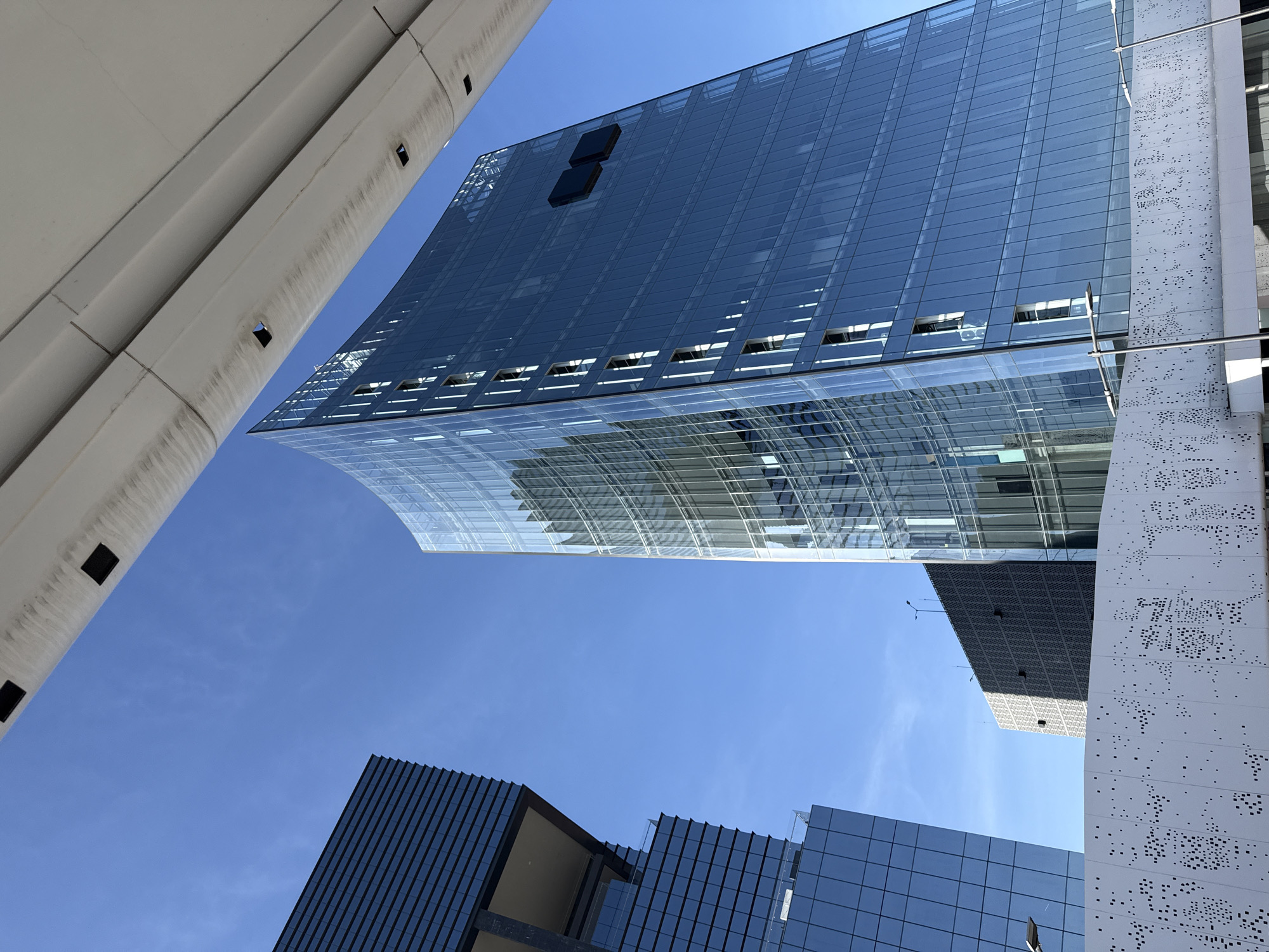 The image is a low-angle shot of modern architecture in Puebla, Mexico. The focus is on several buildings with glass and concrete exteriors. One structure features a curved glass facade and reflective windows. Another building has a perforated white facade, while others display darker glass panels. The sky is clear and blue. No people are visible.