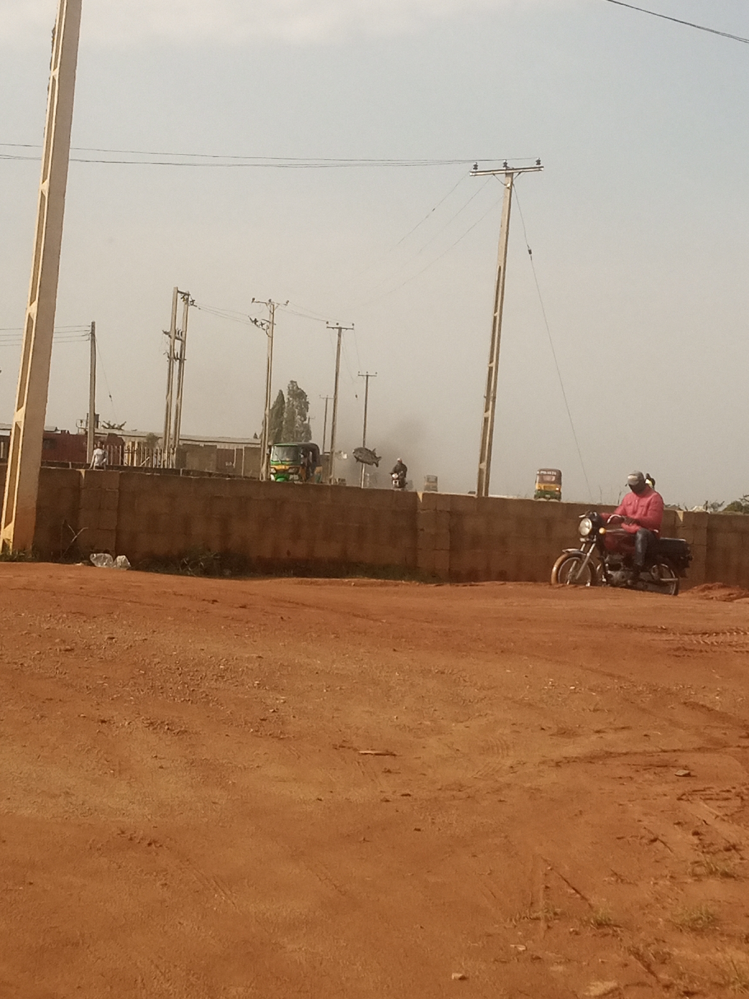 The image depicts a dusty, outdoor scene, likely in Jos, Nigeria, characterized by red earth and a hazy sky. In the foreground, a dirt road or path shows tire tracks, indicating vehicle movement. A large, light-colored concrete pole stands prominently on the left side of the frame.
A low, brown brick wall runs horizontally across the mid-ground, separating the foreground from a slightly elevated area behind it. On the left side of the wall, a person in a white t-shirt is visible standing. Further along the wall, to the right, a green and yellow auto-rickshaw is parked or moving.
Dominating the right side of the image, a person wearing a pink long-sleeved shirt and a cap is riding a dark-colored motorcycle. The motorcycle is partially embedded in the red earth. Another motorcycle is seen further in the background, on the left side, with a rider.
Several utility poles with wires are scattered across the background, suggesting the presence of infrastructure. The overall atmosphere is one of a dry, potentially arid environment, with a subdued color palette.