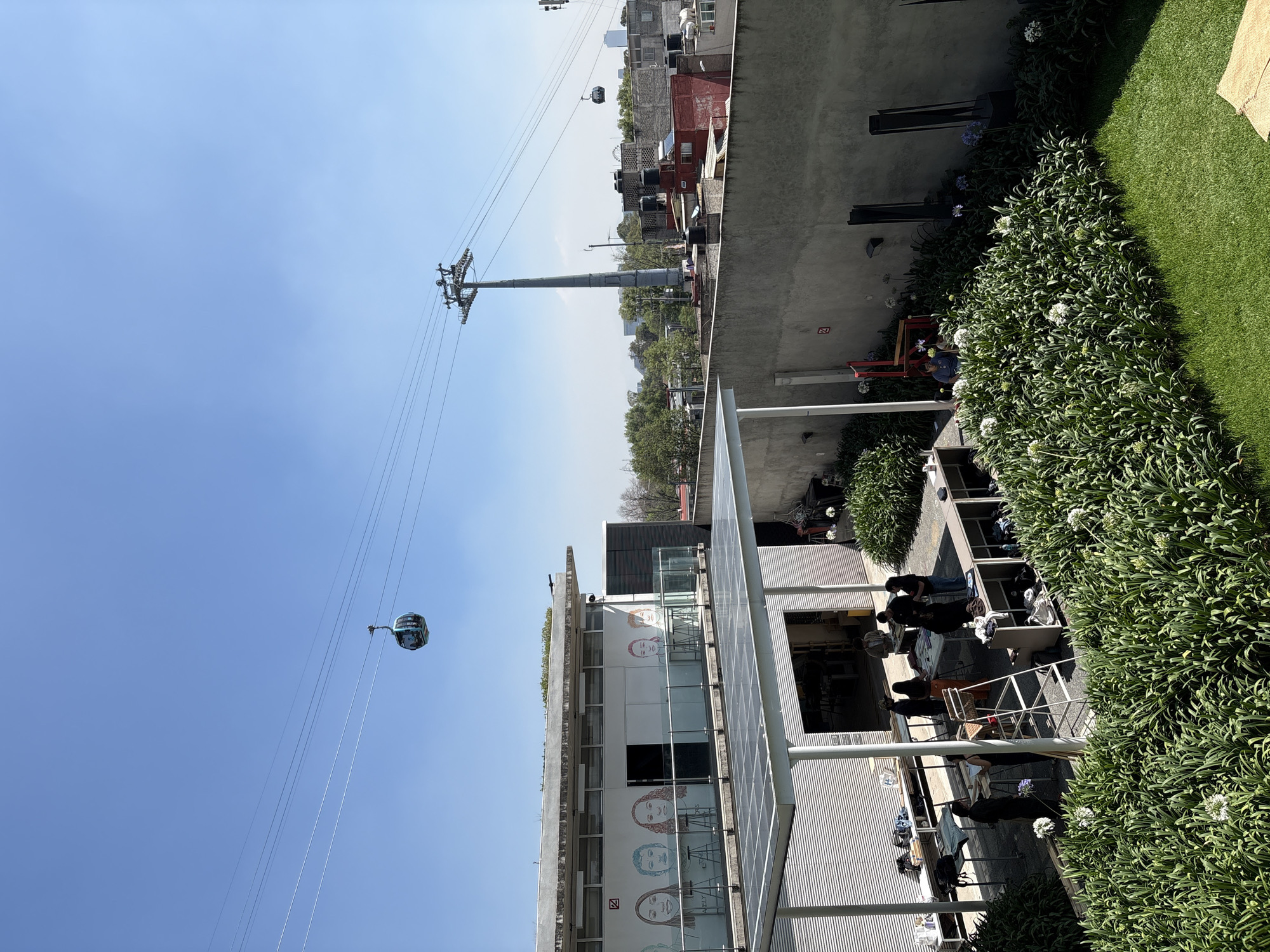 The image depicts a scene in Mexico City, Mexico, with a cable car system prominently displayed against a clear blue sky. The cable car and its supporting tower are visible, with one of the cable car cabins suspended in transit.
In the foreground, there is a building with a modern design, featuring a white facade adorned with portrait illustrations of people. A row of solar panels is installed on the building's exterior. A restaurant or cafe area is situated outside the building, where several people are seated at tables, engaged in conversation and dining. The area is decorated with greenery, including hedges and potted plants.
Beyond the building and the cable car system, the cityscape of Mexico City is visible, with various buildings and trees forming the backdrop. The image is taken during daytime, with bright sunlight casting shadows and highlighting the details of the scene.