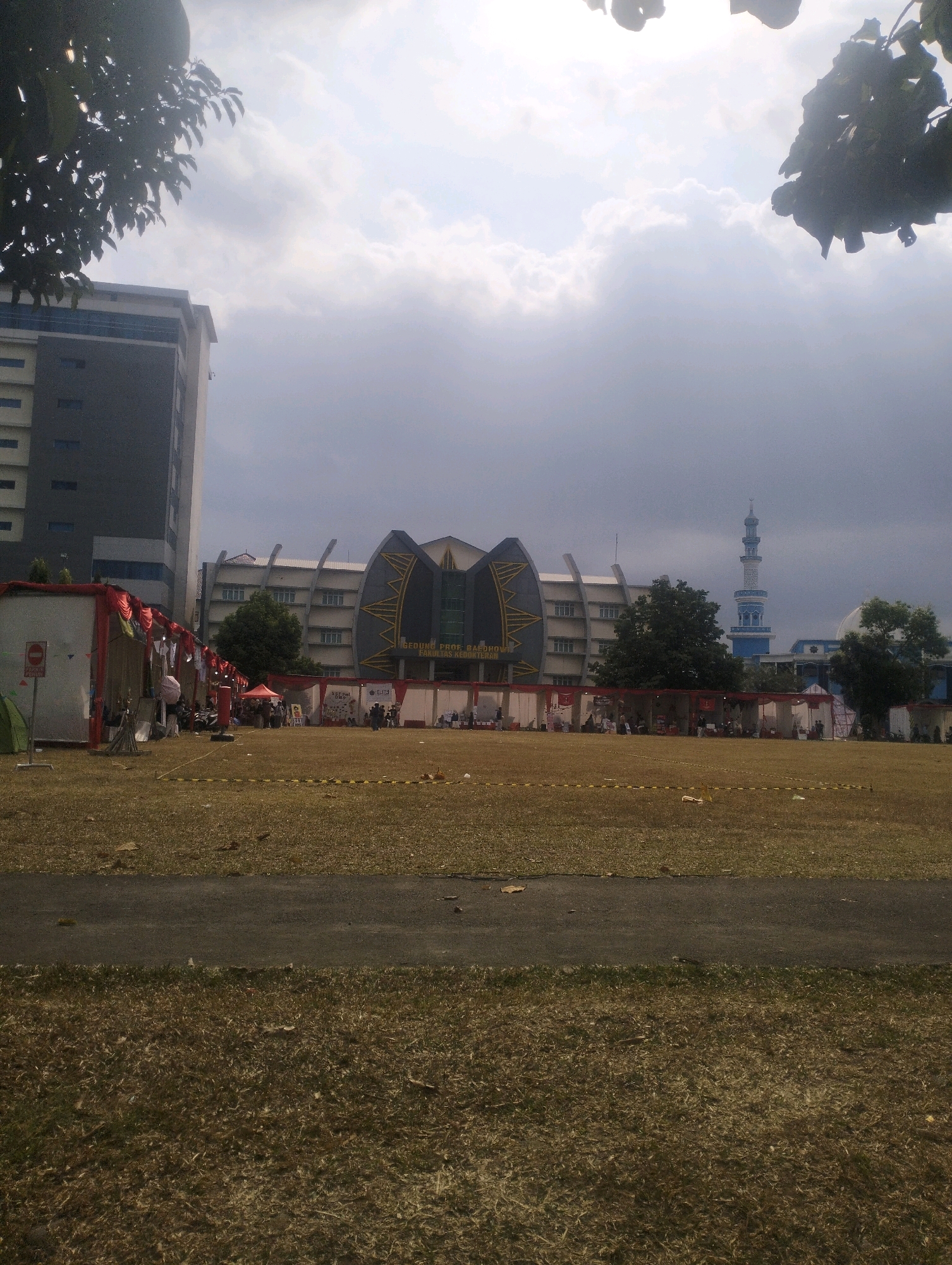 The image depicts a wide outdoor scene on a cloudy day, possibly in Purwokerto, Indonesia. In the background, there are several buildings, including a modern multi-story building on the left, and a larger, distinct building with a unique architectural design in the center. This central building has a sign that appears to read "GEDUNG PROF. RAE... FAKULTAS..." indicating it might be a faculty building. To the right of this building, a tall, slender minaret of a mosque is visible, suggesting a university campus or a mixed-use area.
In the foreground, a large, dry, grassy field occupies the majority of the frame. A paved path runs horizontally across the middle of the field. Along the edge of the field closer to the buildings, there are several white tents or stalls set up, decorated with red fabric and banners. Some people are visible standing and walking around these stalls, and there are a few motorcycles parked behind them. The ground in the field has patches of dry grass and some scattered debris. The overall impression is of an outdoor event or market taking place on the grounds of an educational institution. The sky is overcast with dense clouds.