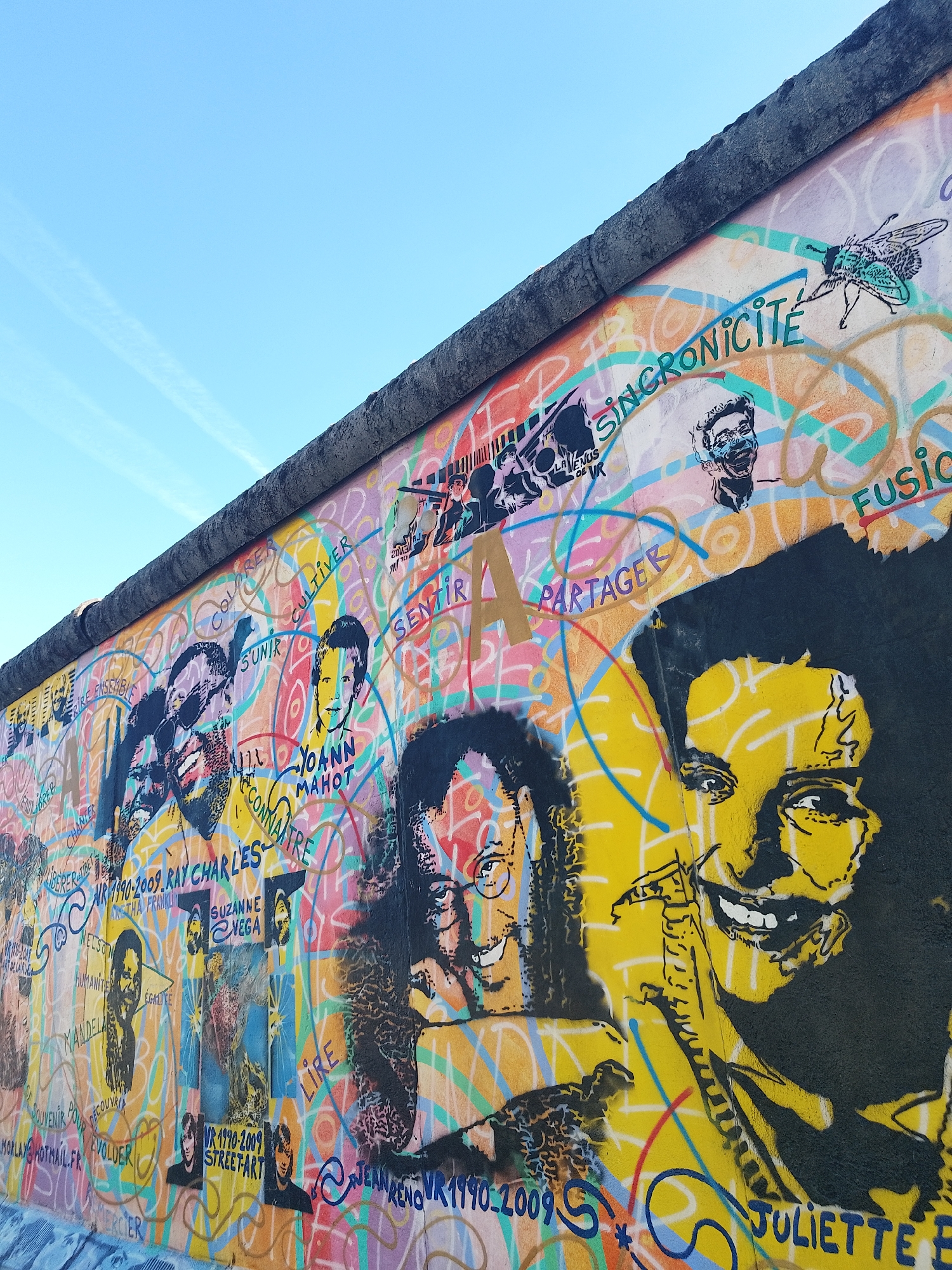 This image shows a section of the East Side Gallery in Berlin, Germany. The visible portion of the Berlin Wall is covered in colorful graffiti and murals, including portraits of famous figures such as Ray Charles, Nelson Mandela, and Juliette Binoche, with accompanying names and years. Other words like "Synchronicity," "Fusion," "Partager," and "Cultiver" are integrated into the artwork. In the upper left corner, there is a patch of blue sky. The wall itself is a gray concrete structure.