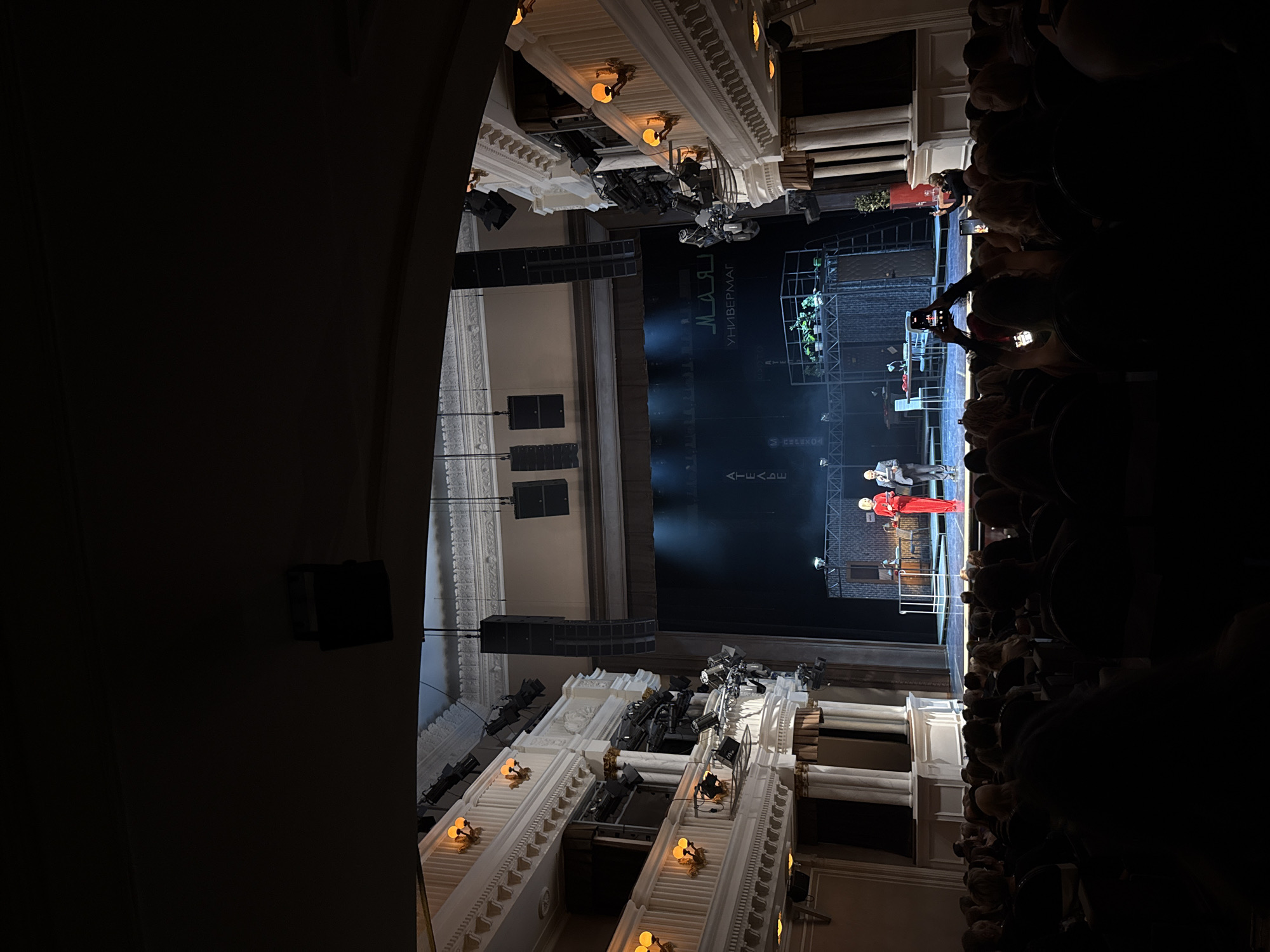 The image depicts a view inside what appears to be a theater. There is a stage with actors in costume performing. A large audience is seated. The architectural details of the theater, including columns, lighting fixtures, and tiers of seating, are visible. Some audience members are holding up phones, possibly to take pictures or videos of the performance. The text on the stage background is in Russian (UNIVERSAL MAE). This is likely the Krasnoyarsk State Opera and Ballet Theatre in Krasnoyarsk, Russia.