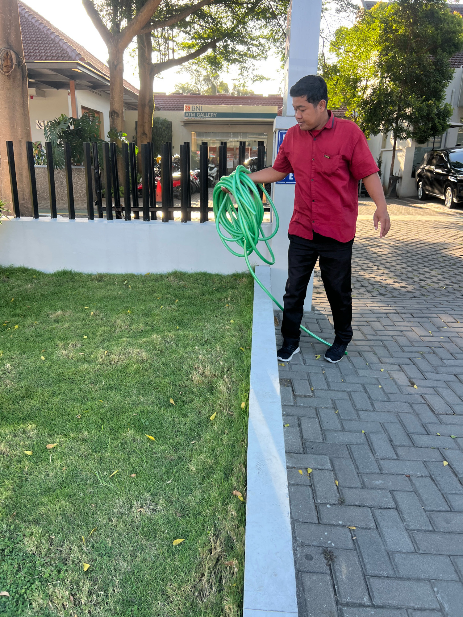 A man in a red collared shirt and black pants is holding a coiled green hose in his right hand. He is standing on a brick pathway next to a patch of grass. Behind him is a white building with a sign that reads "BNI ATM GALLERY". The sun is shining, casting long shadows. The scene appears to be in a public area, possibly near a bank or commercial establishment in Depok, Indonesia.
