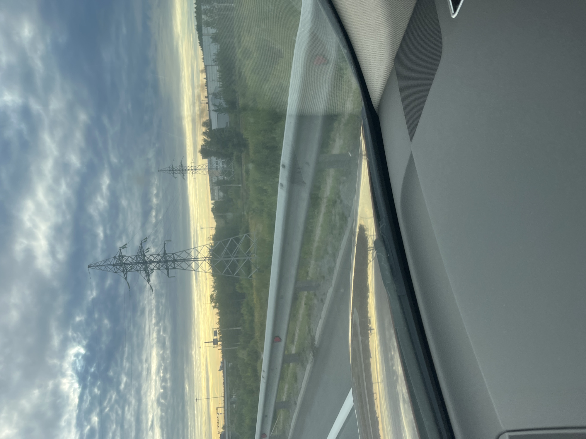 The image captures a view from inside a vehicle on a highway, likely near Saint Petersburg, Russia, given the presence of power lines common in industrial areas and the overcast sky indicative of the region. In the foreground, the dashboard of the vehicle is visible, reflecting the sky and surrounding landscape. A metallic guardrail runs along the side of the highway, with red reflectors attached. Beyond the guardrail, a grassy embankment slopes down, leading to a field dotted with trees and vegetation. In the mid-ground, two large power transmission towers stand prominently against the cloudy sky. The sky itself is a mix of dark, dramatic clouds and lighter patches, suggesting either dawn or dusk. In the background, industrial buildings and more trees are visible, with a plume of smoke rising from one of the structures, hinting at an active industrial zone. The overall scene conveys a sense of transit through an urban fringe area under a moody sky.