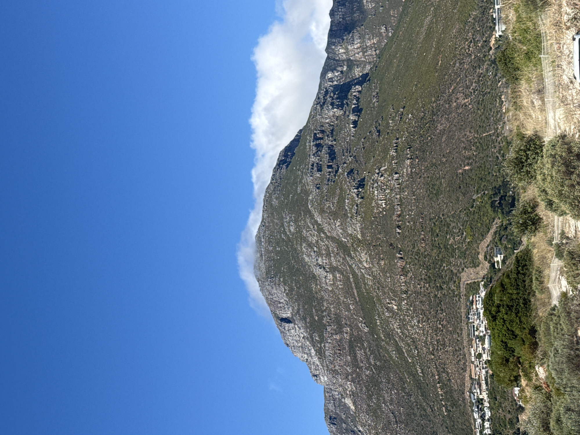 The image presents a view in Hout Bay, South Africa. A mountain occupies the right side of the frame, with its peak shrouded in clouds, and the town sits along the base of it. The sky is clear and blue. The scene features vegetation on the mountain and along its base. The buildings appear to be residential and are white.