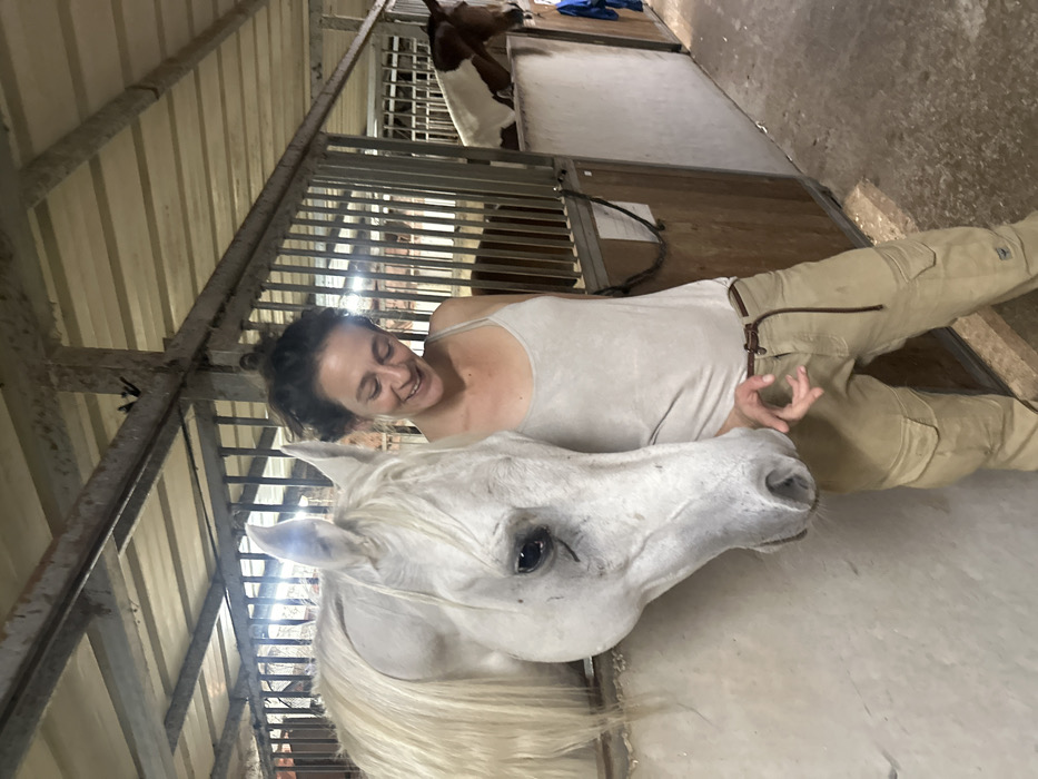 The image depicts an indoor stable in Kfar Monash, Israel. A fair-skinned female with dark hair styled in a bun, wearing a light-colored tank top and khaki cargo pants, stands in the foreground, looking down and to her left with a smile. Her left hand is raised near the muzzle of a light-grey or white horse. This horse's head and neck are visible, extending out of an open stall. The stall features a solid light-colored lower wall and vertical metal bars above. In the background, another horse with a brown and white pinto coat is partially visible within a separate stall, behind metal bars. The stable ceiling consists of light-colored corrugated metal sheets and support beams. The floor is an uneven concrete or packed earth surface. A dark rope hangs on a partition to the right of the female.