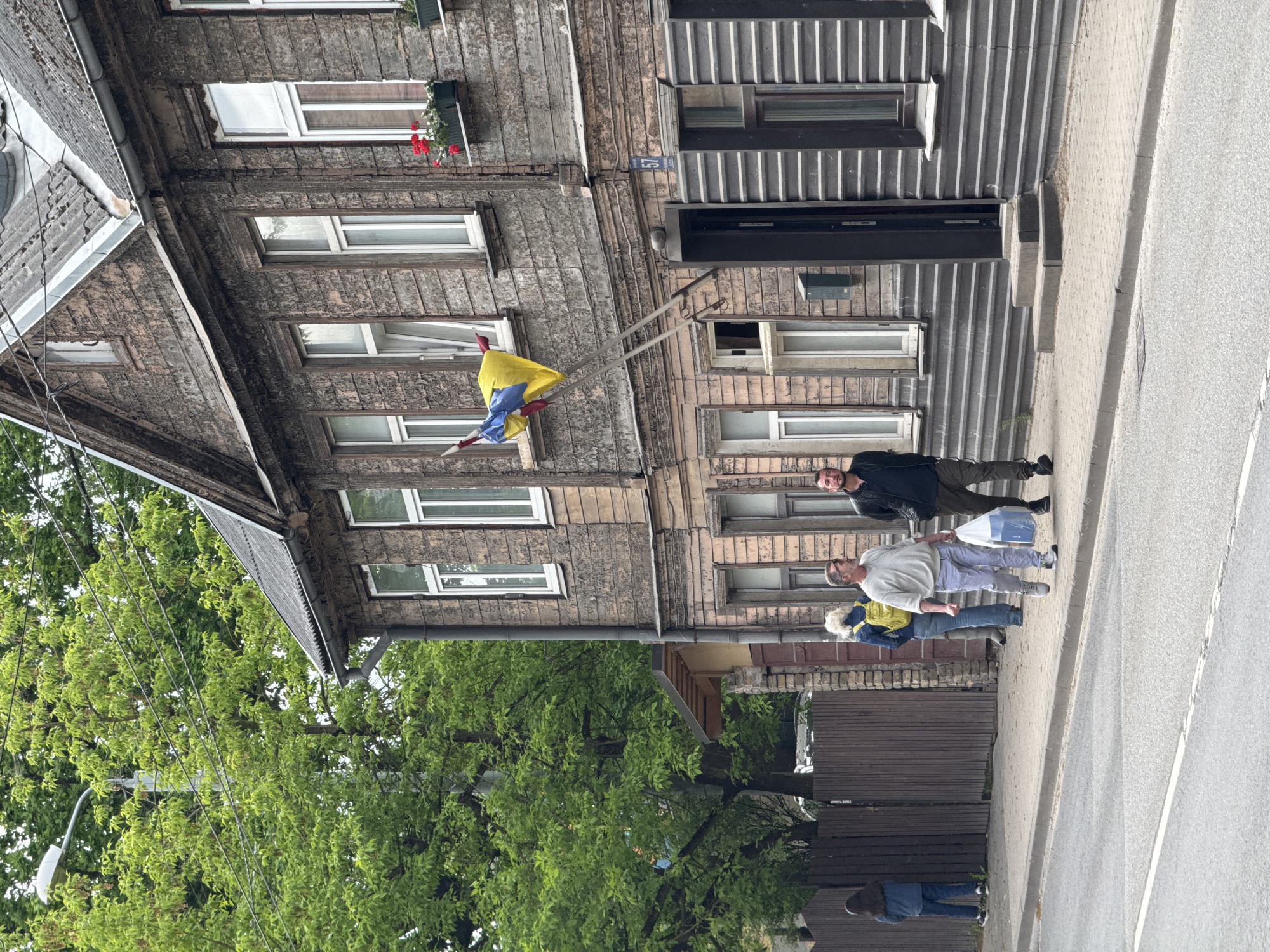 The image depicts a street scene in Riga, Latvia, featuring a weathered, two-story wooden building with multiple windows, some adorned with flower boxes. A flag, displaying yellow, blue, and red stripes, is attached to a pole extending from the building's facade. Several people are walking on the sidewalk adjacent to the building, including a man in a black jacket and khaki trousers, a man in a grey sweater and sweatpants, and another person with a yellow backpack. A tree with green foliage dominates the lower left corner. The street and sidewalk appear typical for a residential area in Riga.
