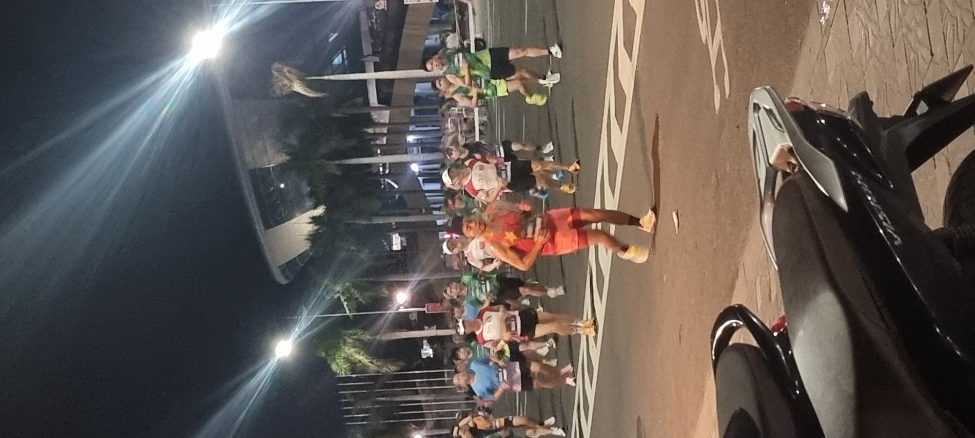 This is a nighttime photograph of a marathon race on a city street in Đà Nẵng, Vietnam. The foreground is dominated by the seats of two parked motorcycles. Beyond the motorcycles, a group of runners are actively participating in the marathon. The runners are wearing athletic attire, including shorts and tank tops, with some wearing race bibs. The road is marked with white lines, and a stadium or large building with a curved roof and palm trees are visible in the background. The lighting appears to be from streetlights and possibly stadium lights, creating bright streaks of light in the dark sky. The overall atmosphere suggests a public event taking place in an urban setting at night.