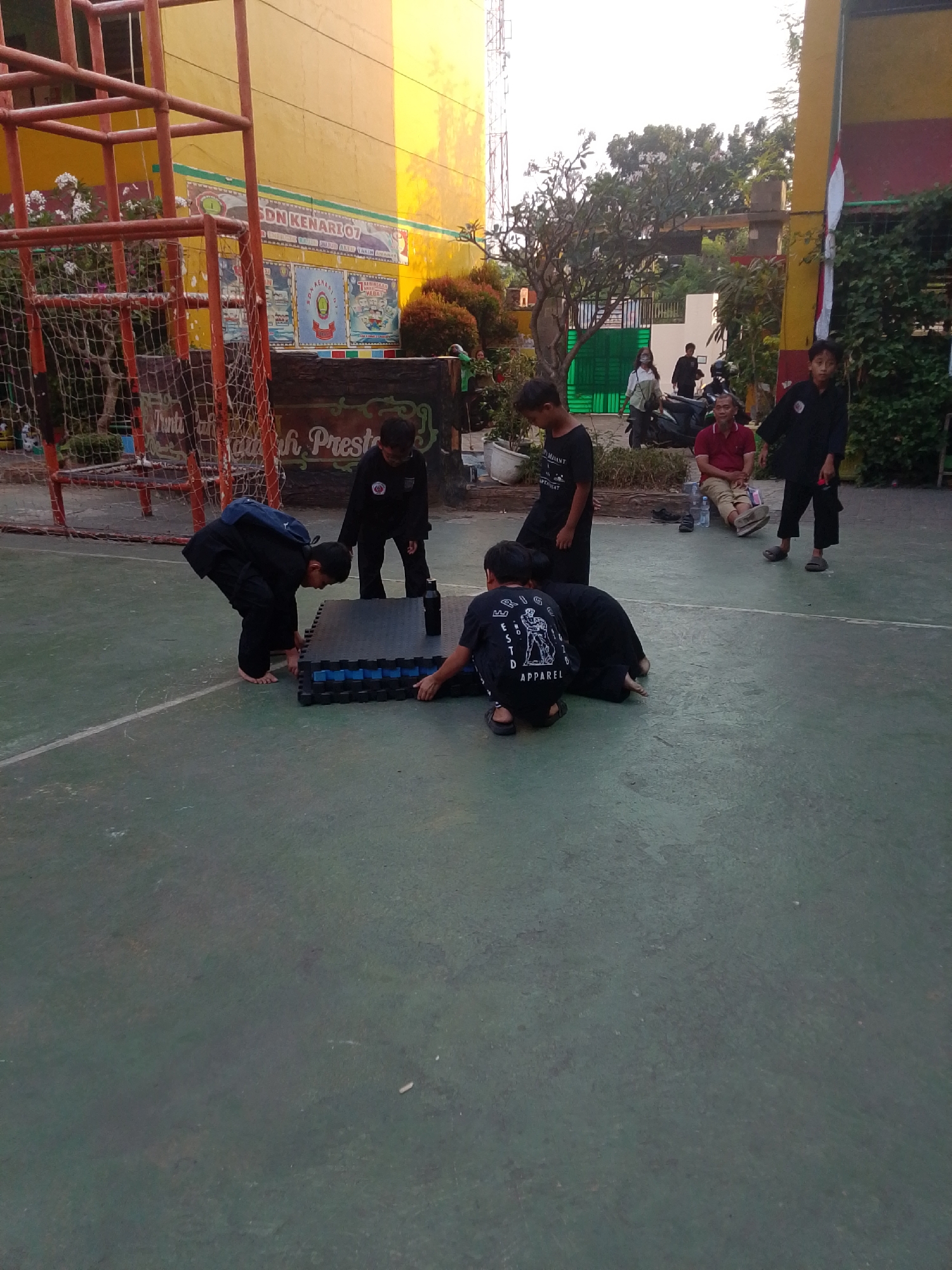 The image depicts an outdoor scene at what appears to be a schoolyard in Central Jakarta, Indonesia. Several children dressed in black martial arts uniforms are interacting with a stack of interlocking mats on a green asphalt surface. In the background, a yellow building with signage and a red metal structure are visible. Other individuals, including an adult in a red shirt, are also present in the scene. The overall environment suggests a casual, everyday activity possibly related to martial arts practice or a school event.