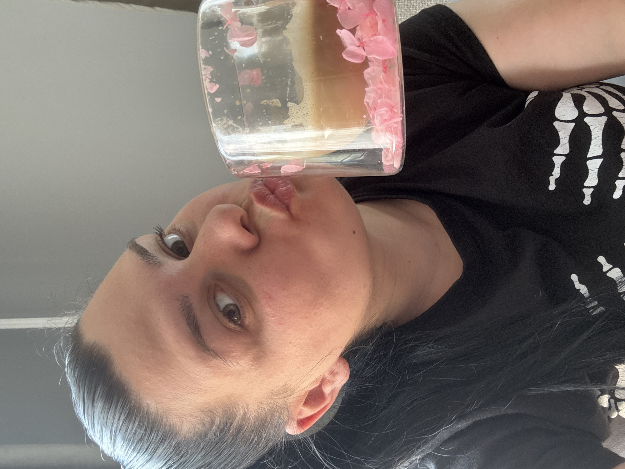 This close-up image, rotated ninety degrees clockwise, features a person (appearing female) with dark hair and dark eyes looking directly at the camera. Their head is tilted, and their lips are pursed in a puckering motion, touching the bottom edge of a clear, square-shaped bottle.
The bottle contains a light brown, slightly frothy liquid, possibly coffee or a similar beverage. Pink, petal-like decorative elements are visible inside the bottle, either floating in the liquid or adhered to the inner surface, creating a distinct aesthetic.
The person is wearing a black top or t-shirt, which features a white graphic print of a skeleton hand on the right side. The background is a plain, light grey or off-white color, suggesting an indoor setting. The lighting is bright and even, indicating daytime. No specific location cues for Rogosh, Bulgaria, are visible within the frame. There is no visible text in the image.