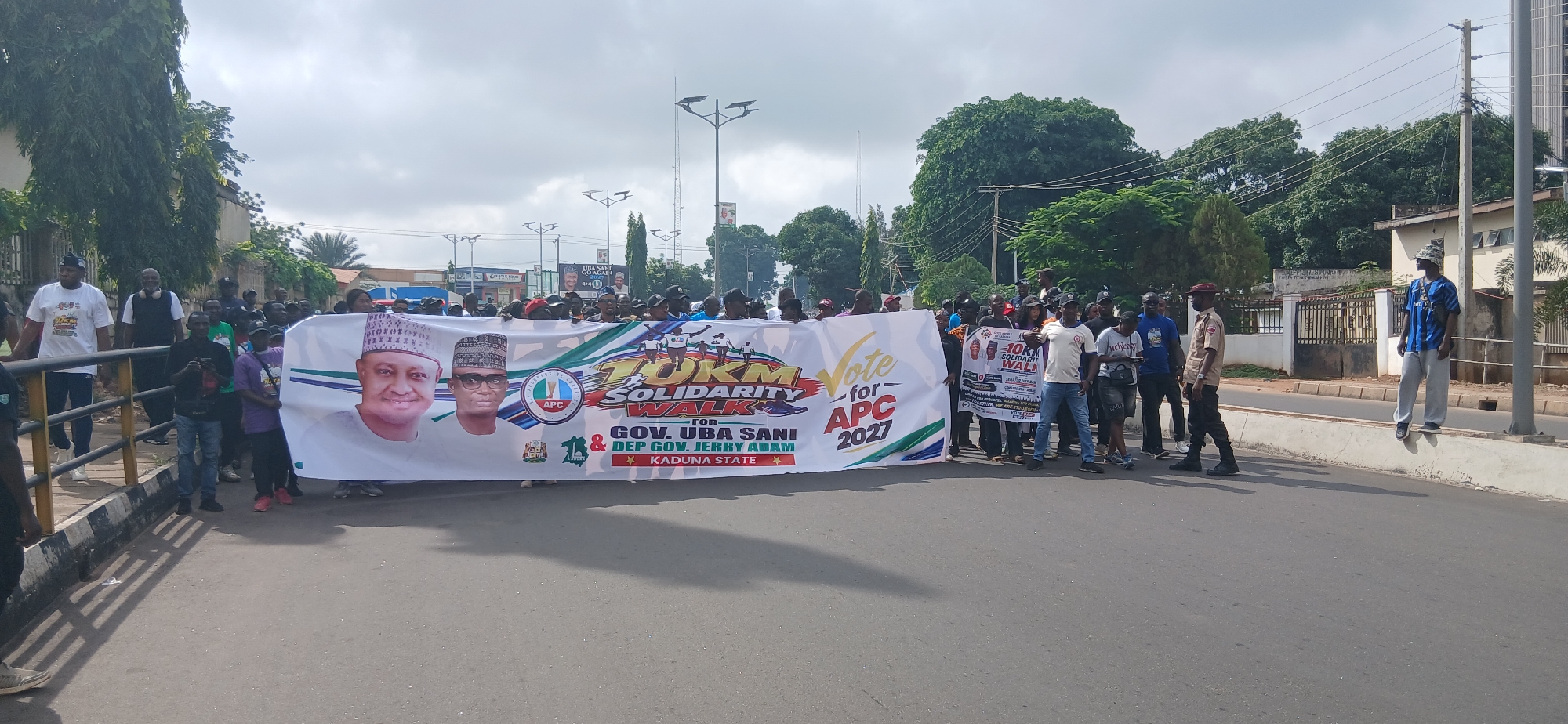 A group of people are walking on a paved road, carrying a large banner. The banner features portraits of two men, one wearing a traditional cap and the other a skullcap, with the text "10KM SOLIDARITY WALK FOR GOV. UBA SANI & DEP GOV. JERRY ADAM, KADUNA STATE" and "Vote for APC 2027". Several individuals are wearing t-shirts, including some with political branding. The environment appears to be a street in a city, with trees and buildings visible in the background, under a cloudy sky. The overall scene depicts a political rally or procession.