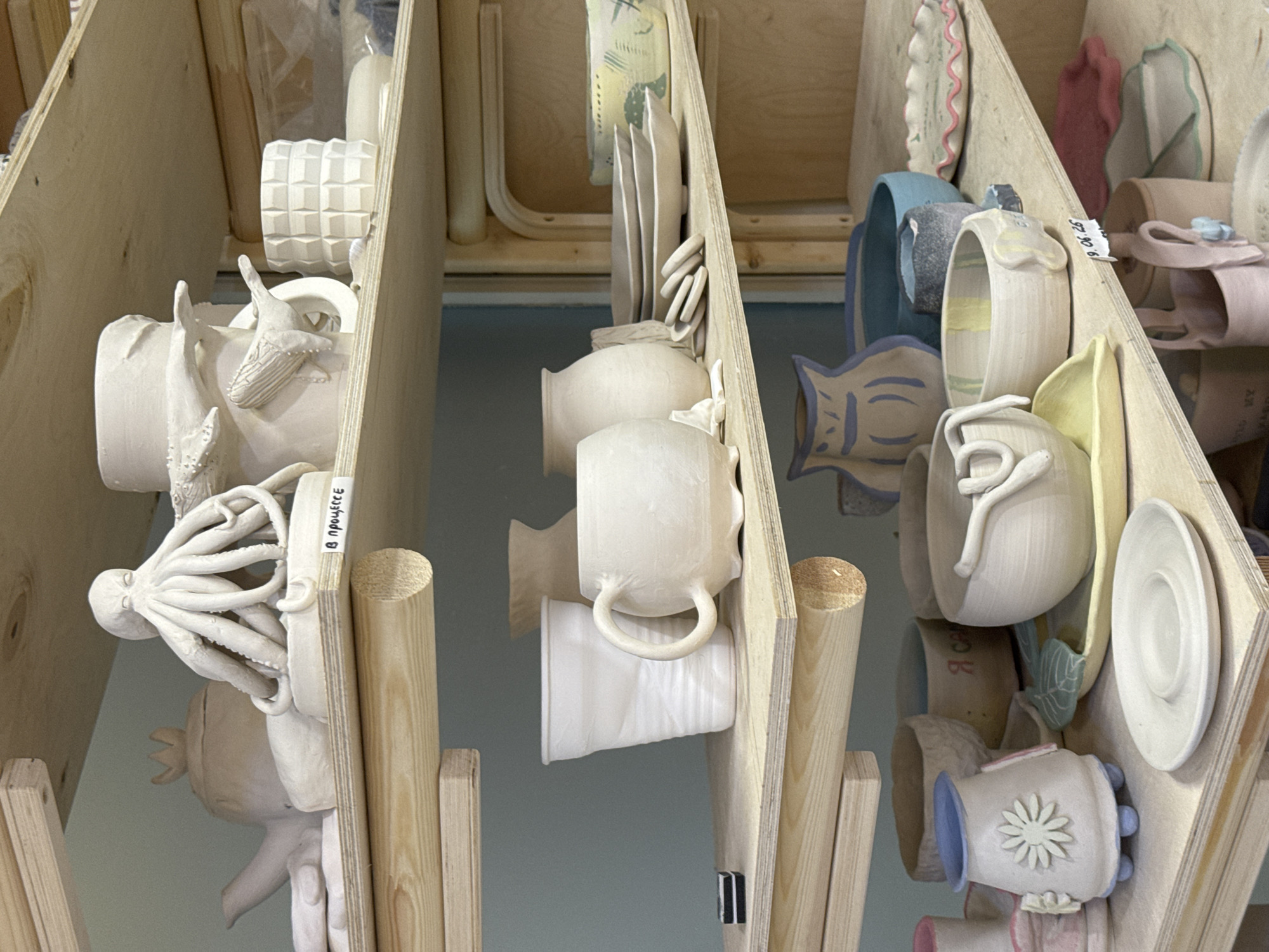 The image depicts a cluttered shelf in what appears to be a ceramics studio. The shelves are made of light-colored wood, and various ceramic pieces are displayed. On the top shelf, there's a teapot with a crown-shaped lid, an octopus sculpture, and a mug decorated with whales. A label that reads "В ПРОЦЕССЕ" (in progress) is visible.
The middle shelf holds several white cups, a larger mug with a handle, stacked plates, and a decorative bowl with green and yellow patterns.
The bottom shelf features more ceramic items, including small vases with floral and blue designs, bowls, and saucers. A small label with handwritten text and "9.06.25" is attached to one of the items.
The overall impression is that of a workspace filled with unfinished and completed ceramic projects. There are no people visible in the image, and the environment appears to be an indoor studio. The specific location of Moscow, Russia, is not indicated by any visual cues within the image itself.