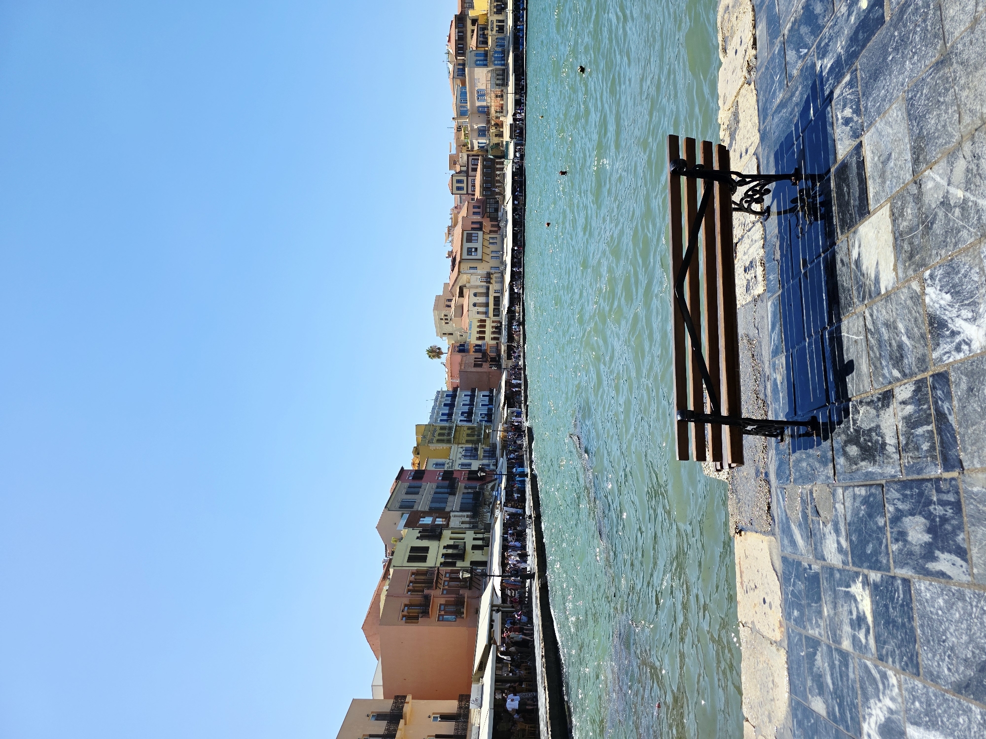 The image depicts the Venetian harbor in Chania, Greece, on a sunny day. In the foreground, a wooden bench with black metal legs sits on a paved walkway. The walkway is made of dark, glossy tiles with white veining, interspersed with lighter stone slabs near the water's edge. The sea is a vibrant turquoise, with gentle waves creating ripples on the surface. In the middle ground, a stone quay lines the harbor, with numerous people walking and dining at outdoor cafes. Behind the quay, a row of colorful buildings, typical of Venetian architecture, stretches along the waterfront. These buildings display various shades of orange, yellow, pink, and blue, with arched windows and balconies. The sky above is a clear, bright blue.