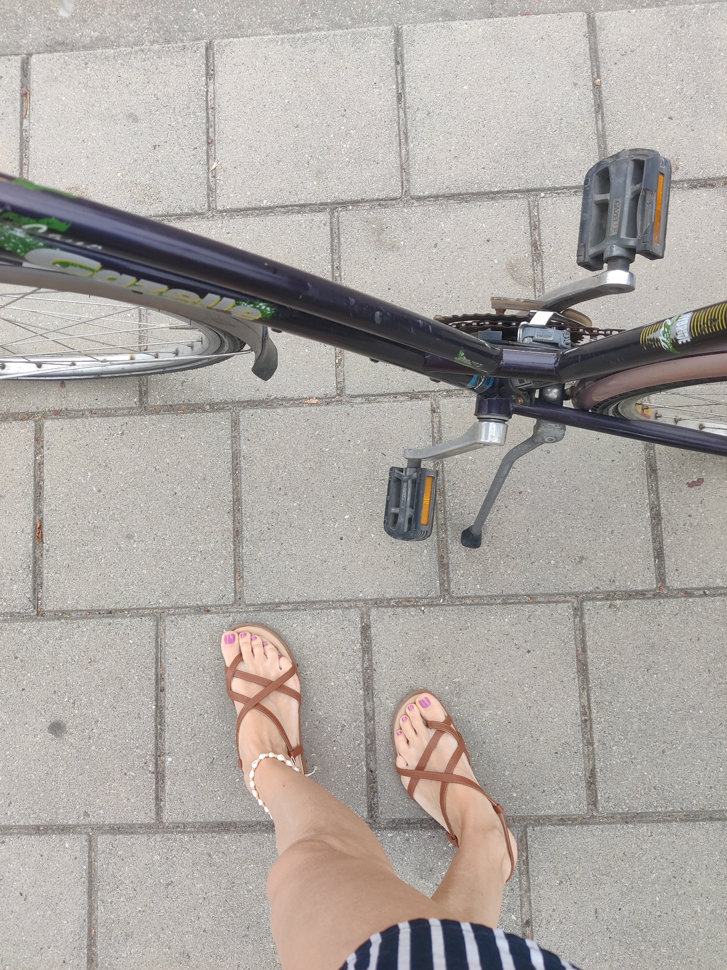 The image, taken in Winterswijk, Netherlands, shows a top-down view of a person's legs and feet, wearing brown cross-strap sandals with a pearl anklet, standing next to a dark purple bicycle. The bicycle's frame has "Gazelle" branding visible. The bicycle is parked on a sidewalk paved with rectangular concrete blocks. The person's toenails are painted purple. The background consists of the concrete sidewalk.