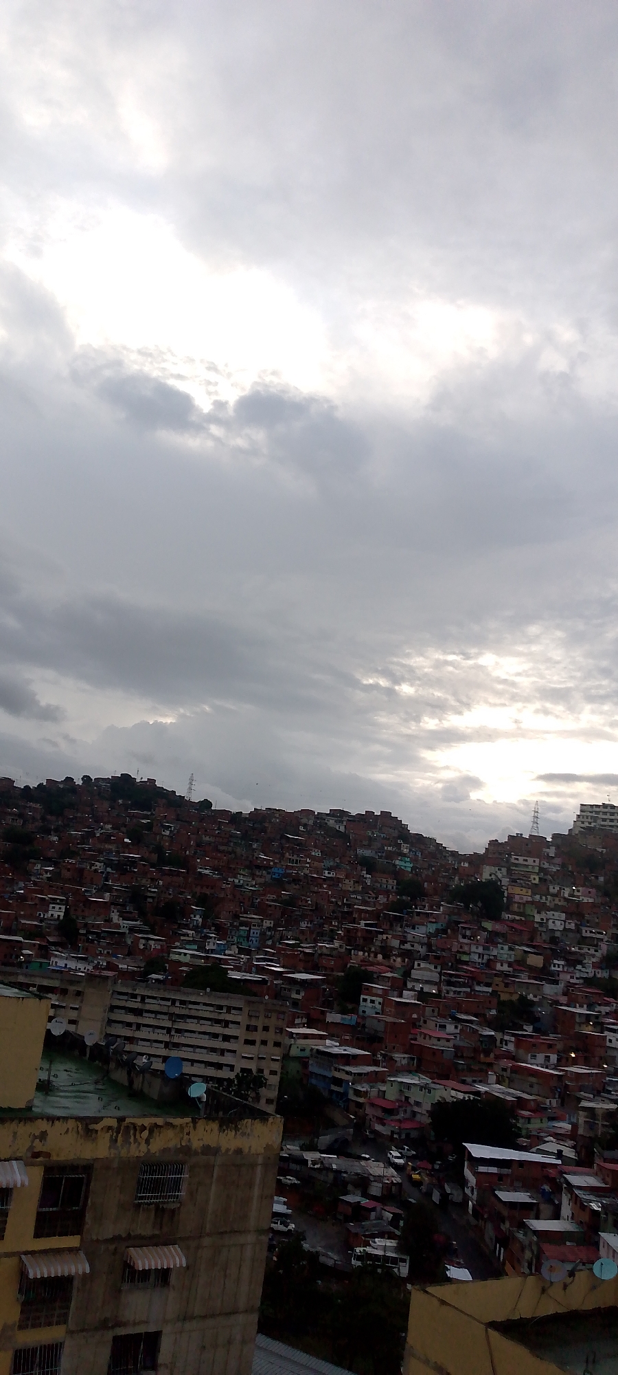 The image captures an aerial view of a dense urban hillside neighborhood in Caracas, Venezuela, under a cloudy sky. The hillside is covered with numerous tightly packed houses, predominantly constructed with red brick and adorned with various colored roofs and facades, giving the impression of a sprawling favela. Some larger, more modern apartment buildings are visible in the foreground and scattered within the hillside, some with satellite dishes on their roofs. The overall impression is one of a densely populated area built on a challenging topography, with evidence of a somewhat dilapidated urban environment. The sky is overcast with diffused light, suggesting either early morning or late afternoon. There are no visible people or specific actions taking place.