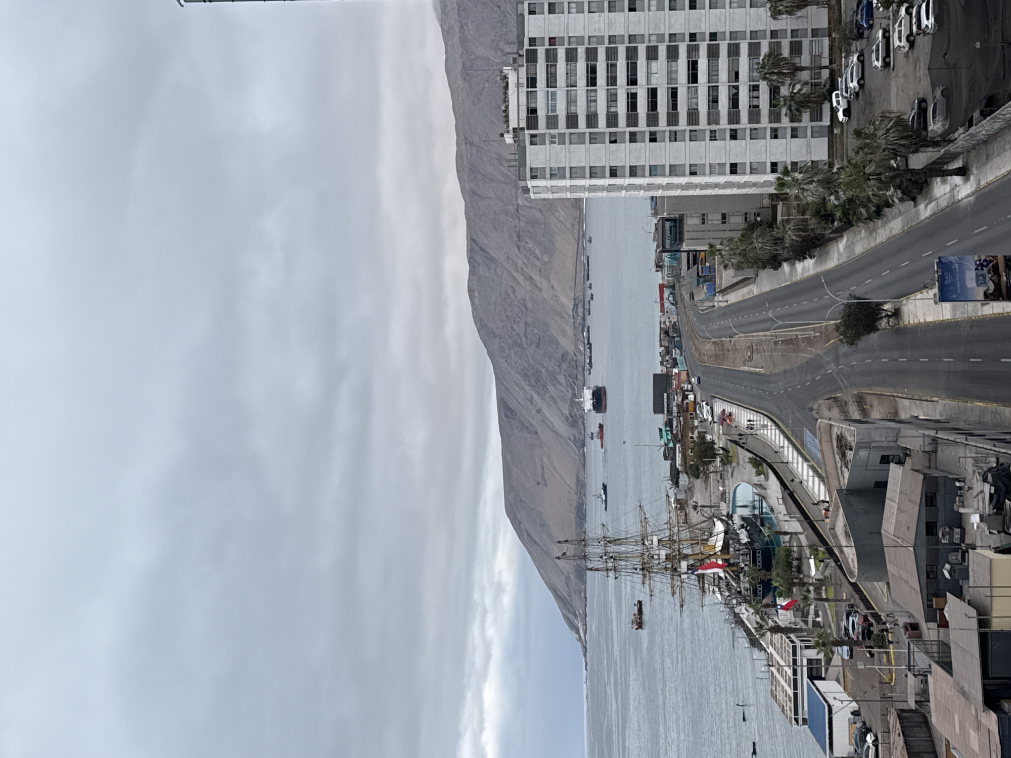 The image depicts a view of the port area in Iquique, Chile, on an overcast day. In the foreground, a multi-lane road runs parallel to the waterfront. To the right of the road is a tall, white, multi-story building with numerous windows. Adjacent to the building is a parking lot filled with vehicles. Further to the left, the waterfront features a docked tall ship, likely a historical vessel, with masts and rigging visible. Several smaller boats and a larger cargo ship are present in the harbor. The sea extends to the horizon, where a large, arid mountain range forms the backdrop. The sky is filled with dense, gray clouds, suggesting a generally cloudy or potentially rainy weather pattern. A few palm trees are interspersed along the road and waterfront. There are no prominent human activities captured in the scene, other than what appears to be a few individuals walking near the docked ship and along the waterfront promenade. The overall impression is one of a coastal urban environment with maritime activity and a dramatic natural landscape.
