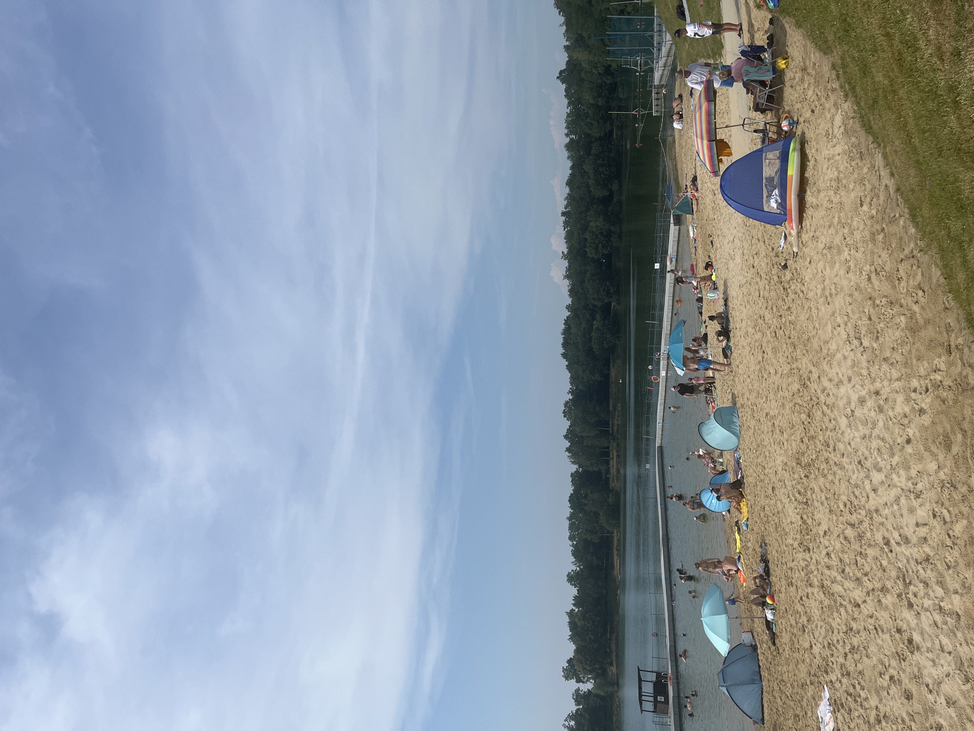 The image depicts a sandy beach area adjacent to a body of water, likely a lake or reservoir, with a treeline in the background. The sky is partly cloudy with a light blue hue. Several people are scattered across the beach, some sunbathing on towels, others seated under umbrellas and tents. A group of people are visible in the water, swimming and wading. There are various beach accessories such as umbrellas, tents, chairs, and inflatable toys present on the sand. The water is calm, and a pontoon or floating structure is visible in the middle distance. The scene suggests a recreational day at a popular outdoor swimming spot.