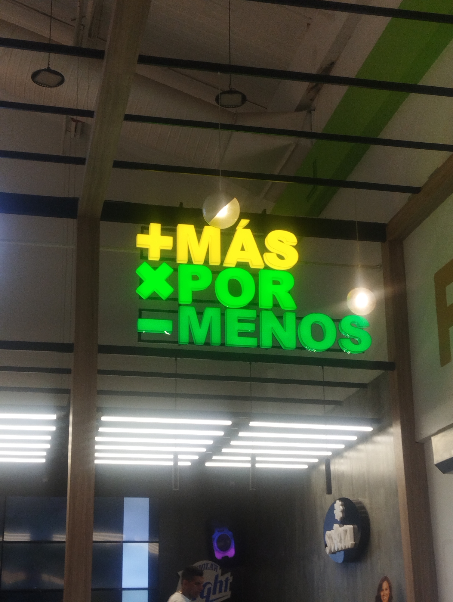 The image shows an interior scene of a retail location in Porlamar, Venezuela. A prominent sign hangs from the ceiling, displaying "+MÁS xPOR -MENOS" in yellow and green neon lettering. The ceiling is exposed, with visible metal beams and lighting fixtures. Below the sign, horizontal fluorescent lights illuminate the interior.
A person is partially visible in the lower-left corner, and a promotional display for "Polar Light" is also present. In the lower-right, a portion of a blue and white logo is attached to a wall, possibly advertising "Swiza." Another person can be seen in the bottom right corner. The overall environment suggests a supermarket or retail store setting.