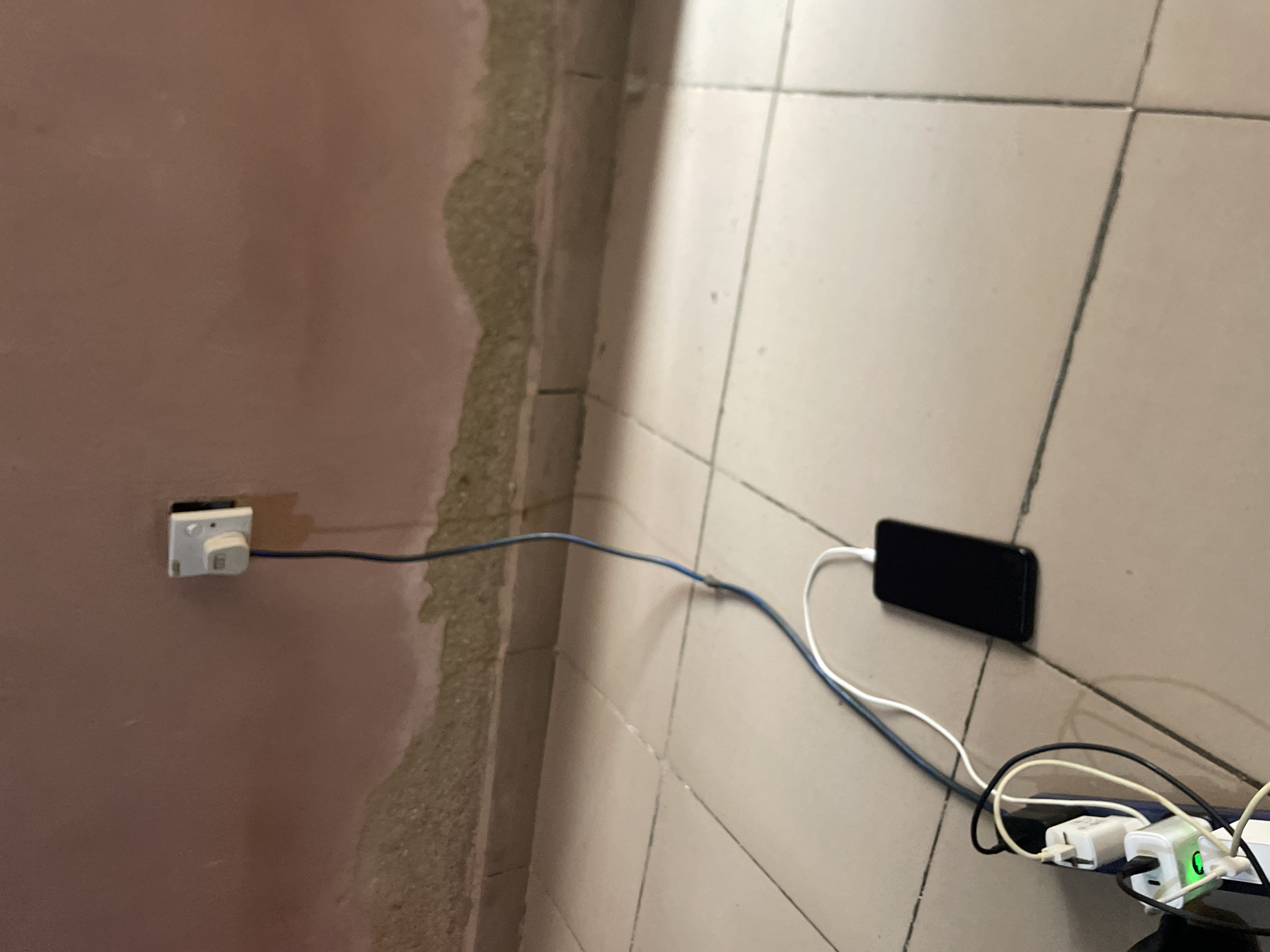 In Jos, Nigeria, a black smartphone is charging on a tiled floor. The phone is connected via a white USB cable to a multi-port charging adapter, which is plugged into a power strip. The power strip is connected to a wall socket by a blue cable. The wall socket is a white, rectangular, three-pin outlet mounted on a beige wall that shows signs of wear and peeling paint. The floor tiles are a light beige color and are laid in a grid pattern. The lighting in the scene is dim, creating shadows on the wall and floor.