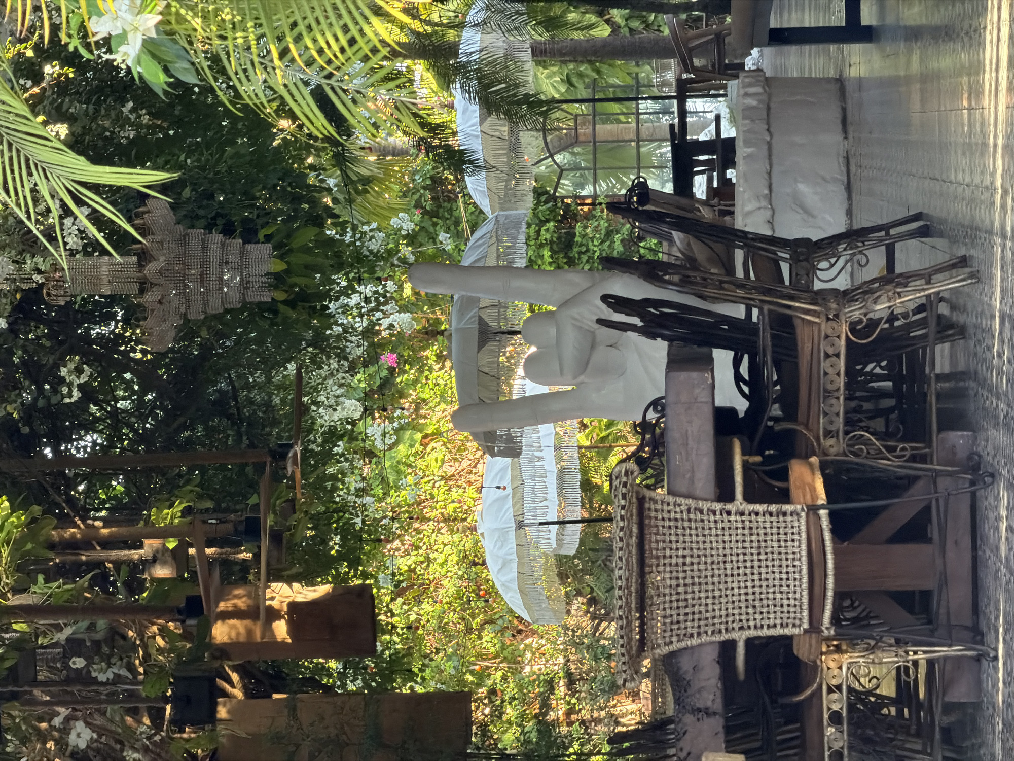 The image shows an outdoor seating area in North Kuta, Indonesia, likely a restaurant or cafe. In the foreground, there is a rustic wooden table with several chairs. One chair is a woven wicker seat with a wooden frame, and others are made of dark metal with intricate scrollwork. Behind the table, a large white sculpture of a hand making a "rock on" gesture is visible, along with two ornate white umbrellas with fringe. Lush green foliage and tropical plants surround the area, creating a vibrant, natural ambiance. A crystal chandelier hangs from the ceiling, adding a touch of elegance. The ground is tiled and reflects the sunlight. The scene suggests a relaxed and stylish outdoor dining experience.