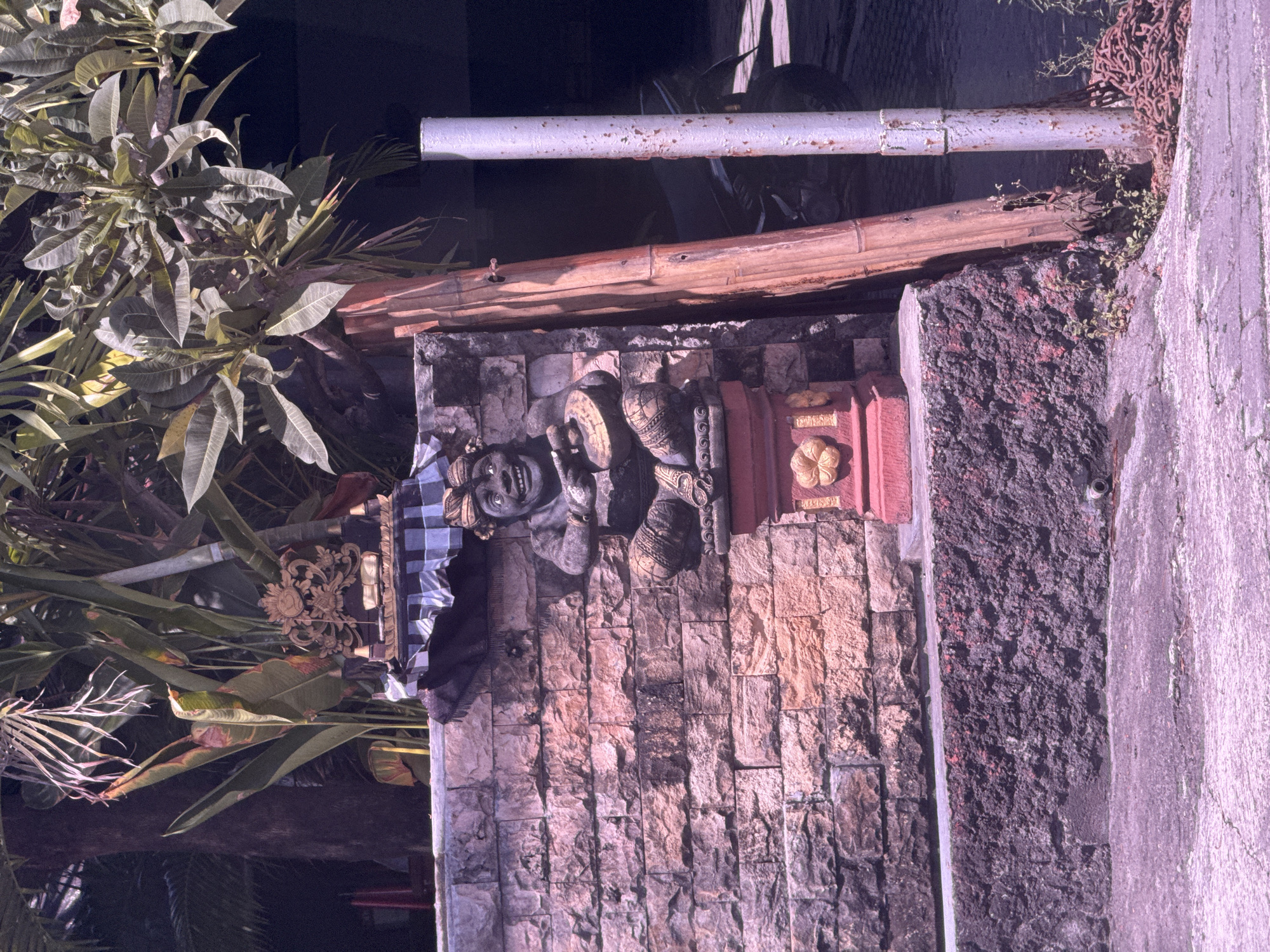 The image shows a stone wall with Balinese carvings and decorations. The wall appears to be part of a traditional structure or gate. There is a stone statue of a human-like figure with a checkered cloth draped on one side. The wall is built of light-colored stones, and there is a decorative element with a flower motif. To the left, foliage from a tropical plant is visible. Above the wall, there are metal pipes and bamboo. To the right of the wall is a rough, dark stone structure with a gray concrete wall next to it. Given the Balinese carvings, it is likely this image was taken in the city of Denpasar, Indonesia.