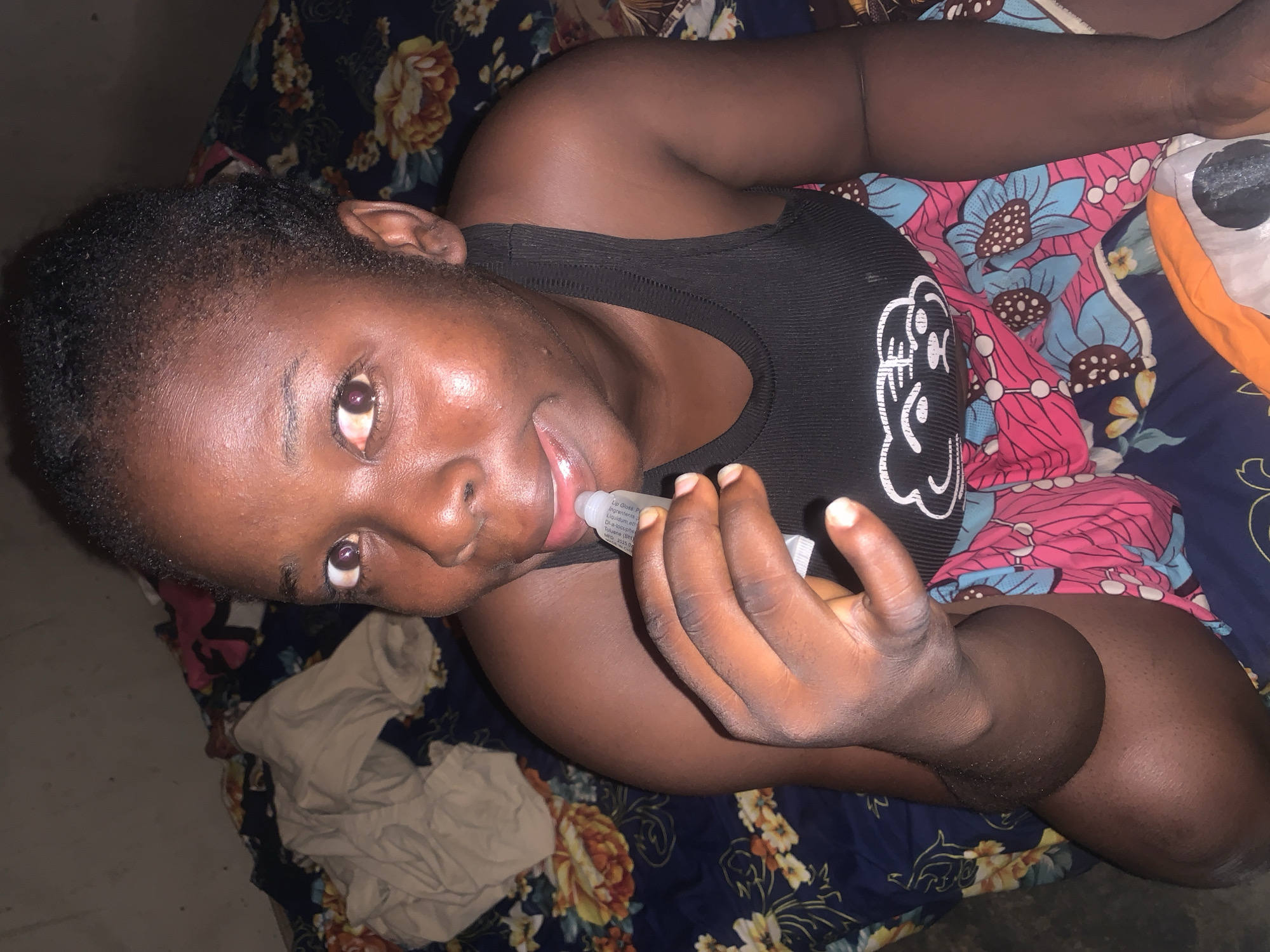 A medium-shot, eye-level indoor photograph captures a dark-skinned woman, positioned on what appears to be a bed with floral-patterned sheets, applying lip gloss. She is wearing a black tank top with a white design on the front. Her gaze is directed toward the camera, and her mouth is slightly open as she applies the lip gloss from a small tube with her right hand. The label on the tube is partially visible, revealing the words "Lip Gloss" and a list of ingredients. The bed is covered in a blue sheet with a floral pattern in shades of brown, orange, and yellow. Underneath this sheet, there is a second layer of fabric with a pink and blue floral pattern. A crumpled white garment is also visible on the bed. The location is presumed to be a residential setting in Agban, Nigeria, based on the instructions.