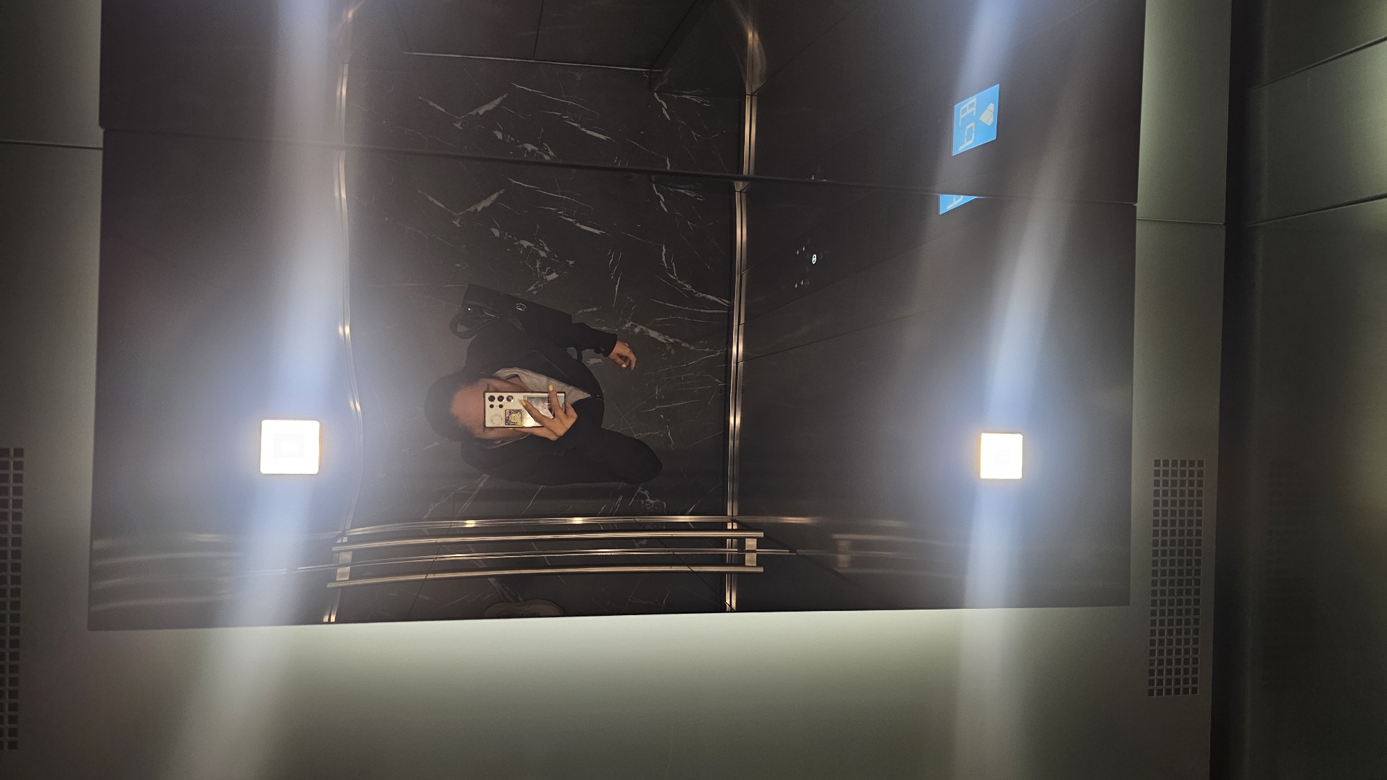 A medium, high-angle shot shows a person taking a picture of themself in a modern elevator, likely in Puebla, Mexico, given the phone's location settings. The elevator has dark gray, marble-patterned walls and shiny metal accents. The subject is wearing a dark blazer, and has a phone with a distinctive camera array. There are modern lighting fixtures and elevator buttons visible.