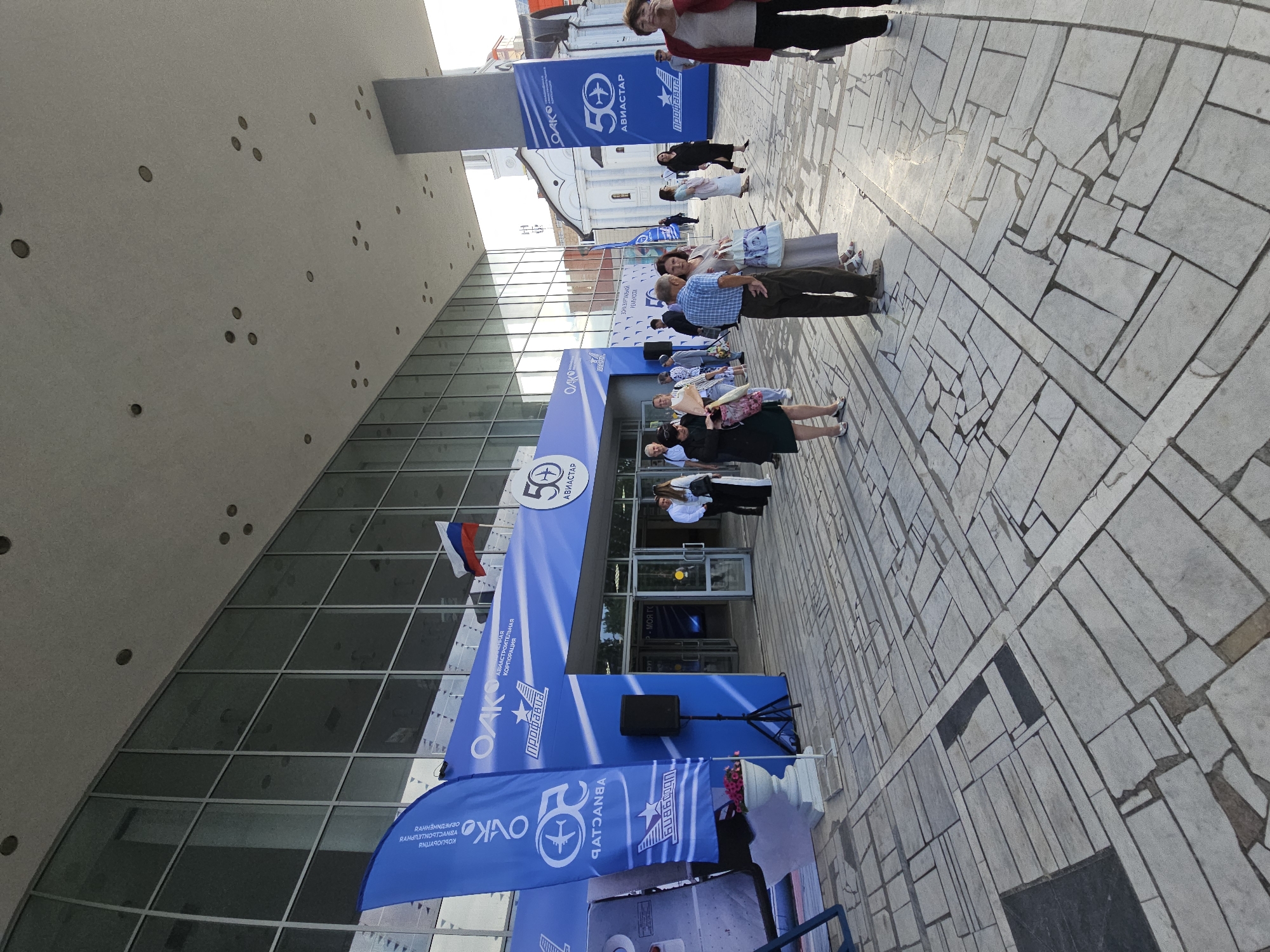 The image depicts an outdoor scene in front of a modern building with a glass facade. Several people are gathered outside, some standing in groups and others walking. The scene appears to be an event, possibly an anniversary celebration, given the prominent blue banners with "50 АВИАСТАР" (50 AVIATAR) and "OAKO" (United Aircraft Corporation) printed on them. One banner also displays the logo of "Профавиа" (Profavia). A Russian flag is visible flying from a pole. The ground is paved with large, irregular light-colored stone tiles. The time of day appears to be late afternoon, with the sun casting some shadows. The overall mood seems festive and organized, suggesting a formal gathering or public event. The building's architecture is contemporary, with clean lines and expansive glass windows reflecting the sky and surrounding environment. Some of the people are dressed casually, while others are in more formal attire.
