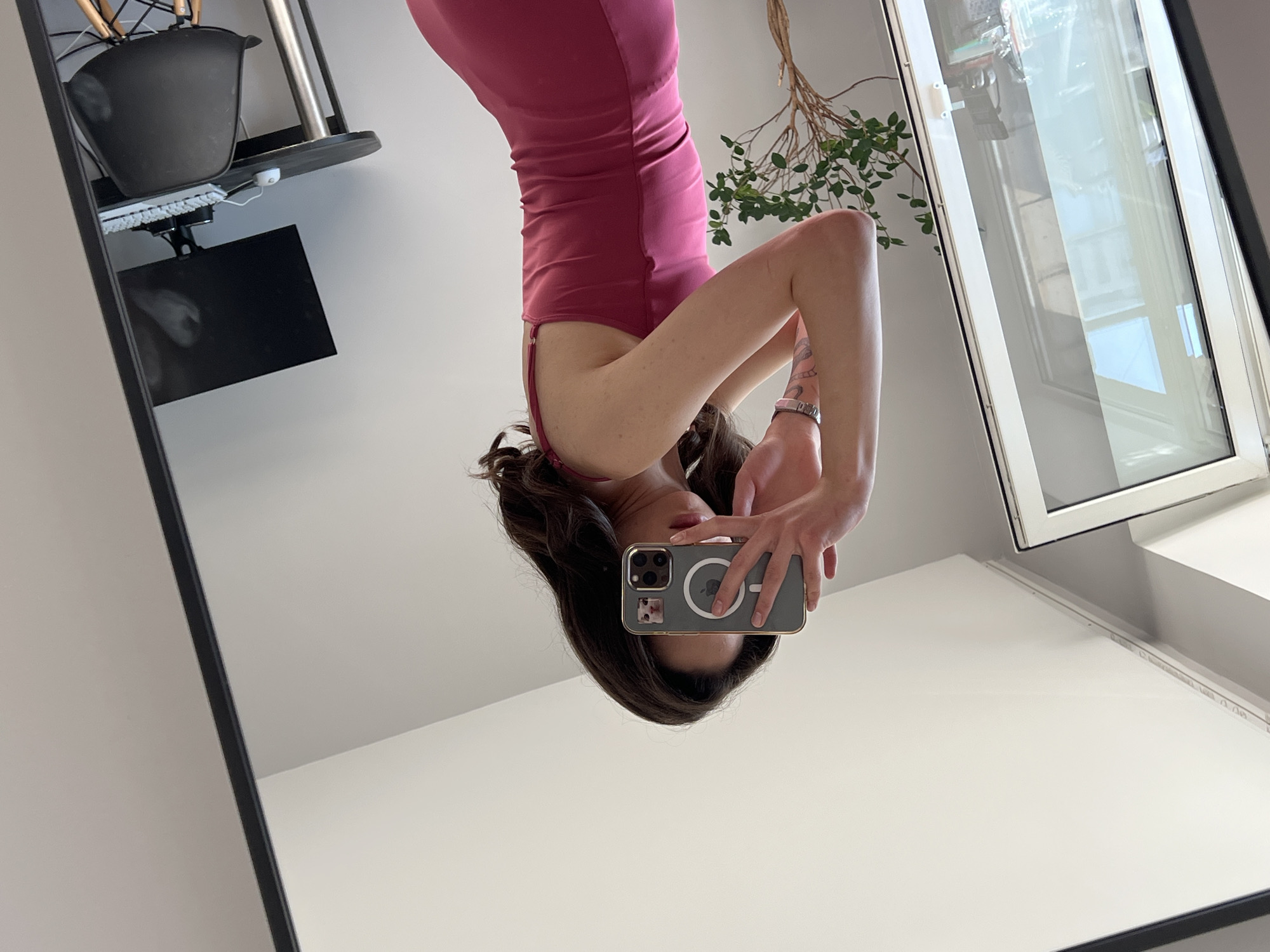 The image captures a woman taking a mirror selfie in what appears to be an apartment. She is wearing a pink slip dress and holding a smartphone in front of her face, obscuring her features. The reflection shows a room with a large mirror, a window with vertical blinds, a desk with a computer monitor, keyboard, and mouse, and a black chair. A small potted tree is visible to the left of the woman. The overall setting suggests a contemporary indoor environment. There is no visible indication of the city of Ternopil, Ukraine.