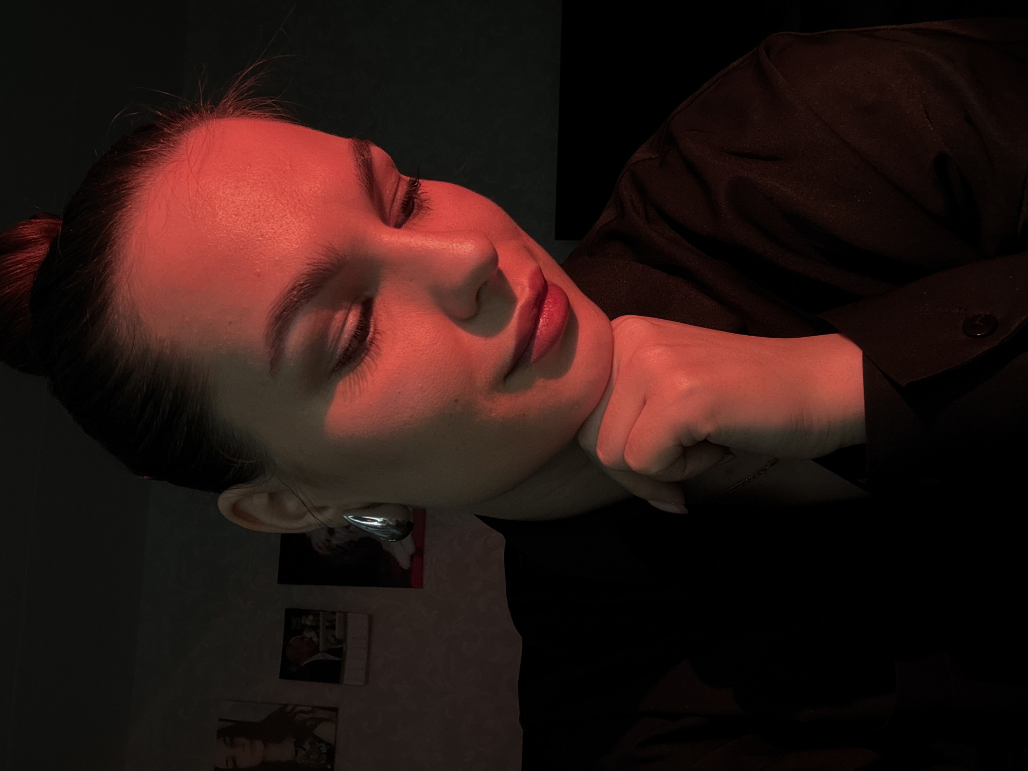 The image is a close-up, medium shot of a young woman from the chest up, positioned slightly off-center to the right. She has her hair pulled up in a bun and is wearing a black collared shirt. Her right hand is resting on her chin, with her thumb and index finger extended. The lighting is dramatic, with a strong red hue casting shadows across her face and upper body. A large, teardrop-shaped silver earring is visible on her left ear. In the background, out of focus, are several framed pictures on a wall. The overall mood is intimate and contemplative. The scene is indoors, with a dark, plain background. There is no discernible indication of the specific location in Chaikovsky, Russia.
