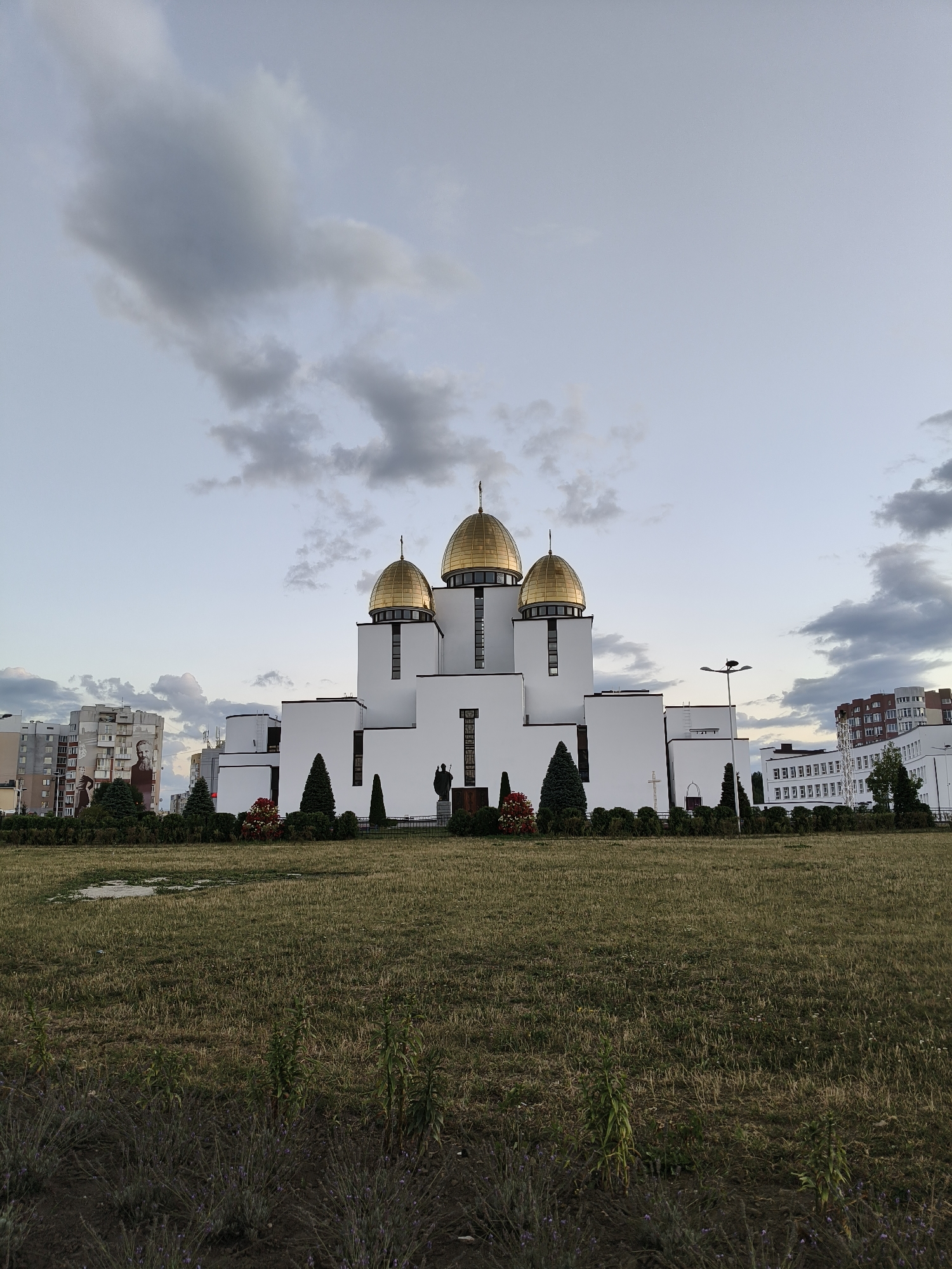 The image shows the Church of the Holy Archangel Michael in Lviv, Ukraine. The church is a modern building with white walls and golden domes. It is surrounded by a lawn and some trees. In the background, there are several apartment buildings and another white building with many windows. The sky is cloudy and the light suggests it is either early morning or late evening. There are no people visible in the image.