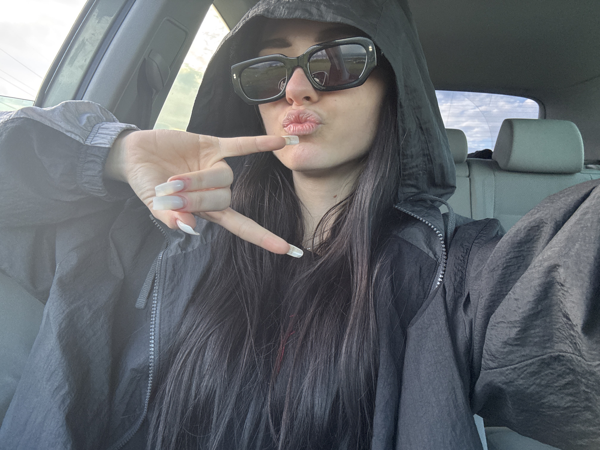 A young woman is in a car, likely the driver's seat. She wears large black sunglasses and a dark hooded jacket. Her hair is long, dark, and straight. She is holding her hand up, making a kissing gesture. The interior of the car appears gray, and outside the back window is a view of a cloudy sky. It is impossible to confirm the location is Burgas, Bulgaria from the provided image.