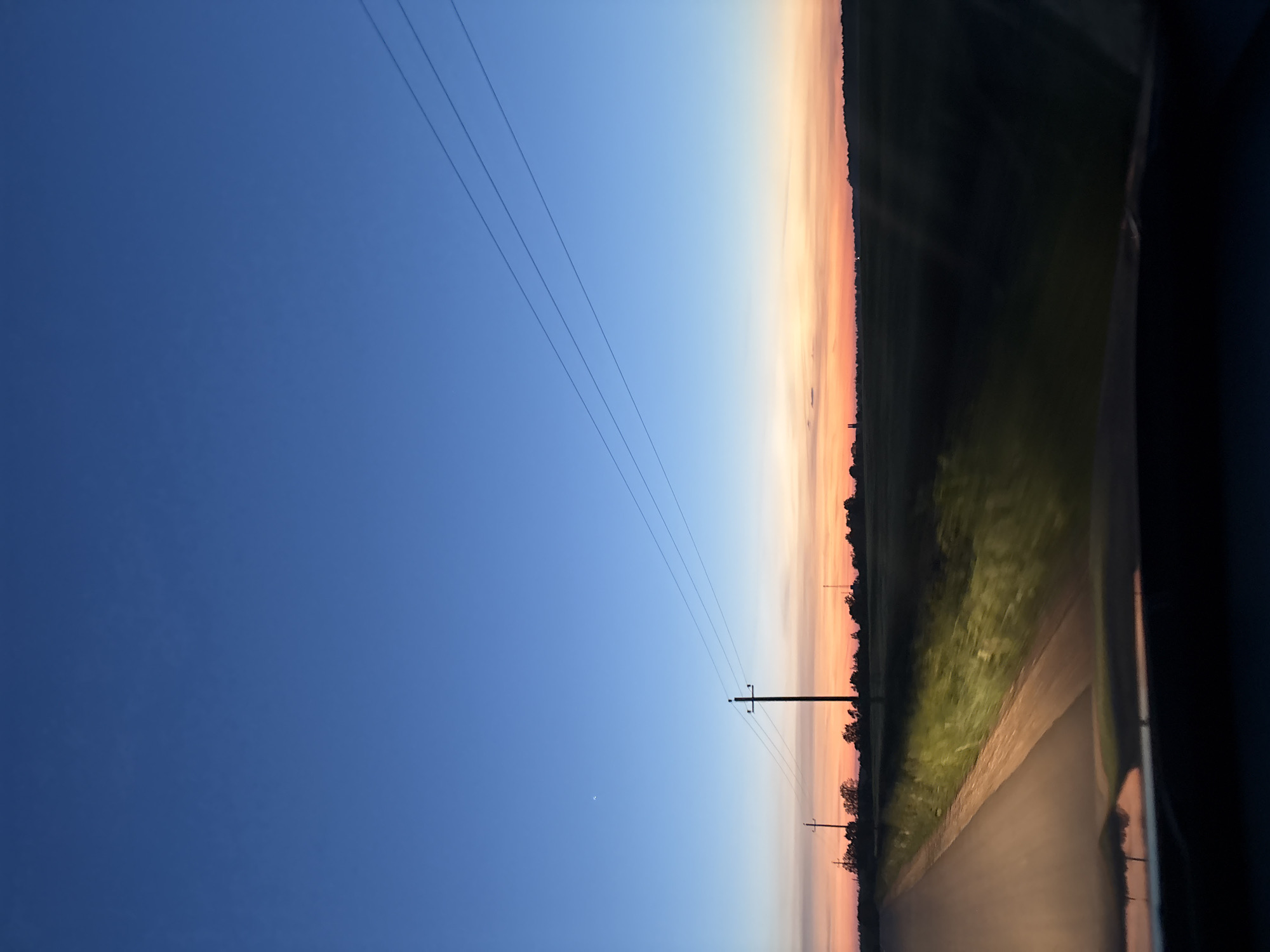 The image depicts an evening scene in Пярэжырскі сельскі Савет, Belarus. A road stretches from bottom right to the vanishing point, flanked by grass and trees on one side. A dark silhouette, likely part of a vehicle's interior, appears on the right edge. Power lines stretch across the upper left part of the image. The sky transitions from a deep blue to a gradient of orange and yellow near the horizon. A single pole and wires are visible in front of the sky.