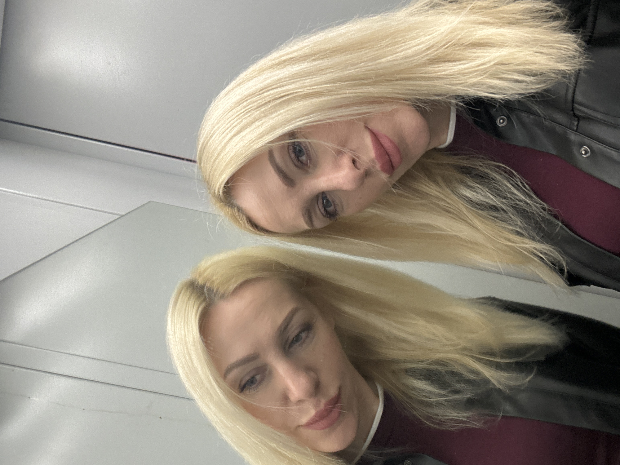 A woman with blonde hair is taking a selfie in what appears to be a fitting room or changing area, as indicated by the mirrored surfaces. She is wearing a maroon top under a black leather jacket. Her reflection is visible in the mirror to her left. The background is a light grey wall with vertical paneling.