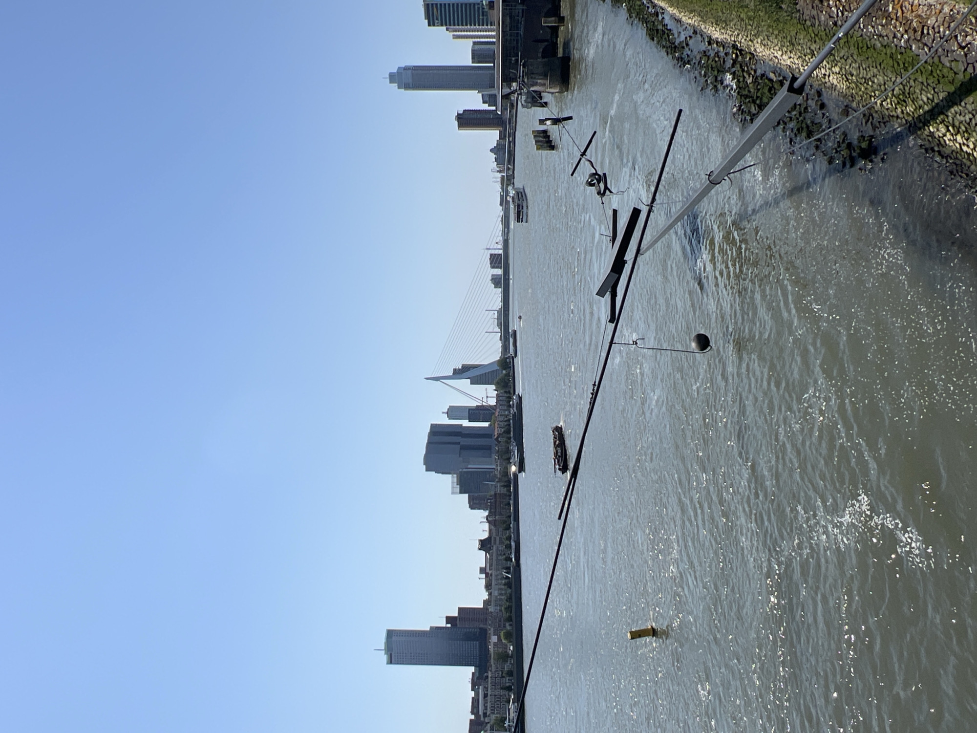 The image shows a panoramic view of the Rotterdam skyline and the Maas River, oriented vertically. The sky is clear and blue, suggesting it is daytime. The sun reflects off the water, creating sparkling highlights.
The river occupies a significant portion of the frame, with several boats visible on its surface. The right bank of the river is lined with a stone embankment covered in green moss and grass. Structures extend from the embankment into the water, possibly parts of a pier or docking facilities, and a prominent long metal pole runs diagonally across the water, with attached wires and a spherical object. A yellow buoy is also visible in the water.
The Rotterdam skyline is visible in the background, a dense collection of modern skyscrapers and buildings of varying heights. A suspension bridge with slender cables stretches across the river, connecting different parts of the city. The architecture of the buildings suggests a contemporary urban setting.
The overall scene depicts a serene day in a bustling port city, with the river acting as a central element of the urban landscape. There is no discernible text in the image.
