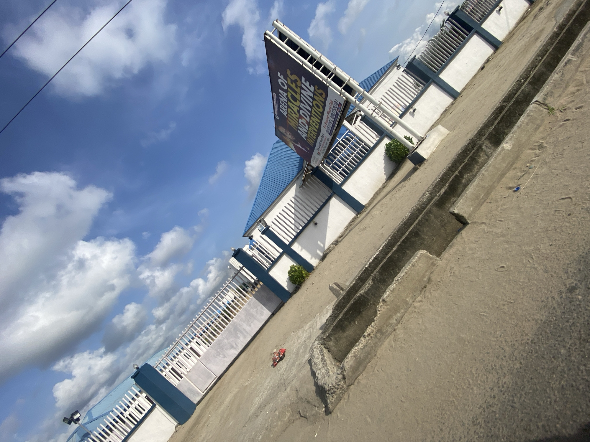 The image, taken at an oblique angle, shows the building of "Arena of Miracles and Divine Interventions," located in Ado, Nigeria, under a partly cloudy sky. The building has a blue roof and white walls with blue trim and is surrounded by a white metal fence with blue posts. A large sign advertising the church is prominently displayed. The foreground features a concrete sidewalk with a drainage ditch. Trash can be seen in the sidewalk. Telephone lines can be seen in the distance.