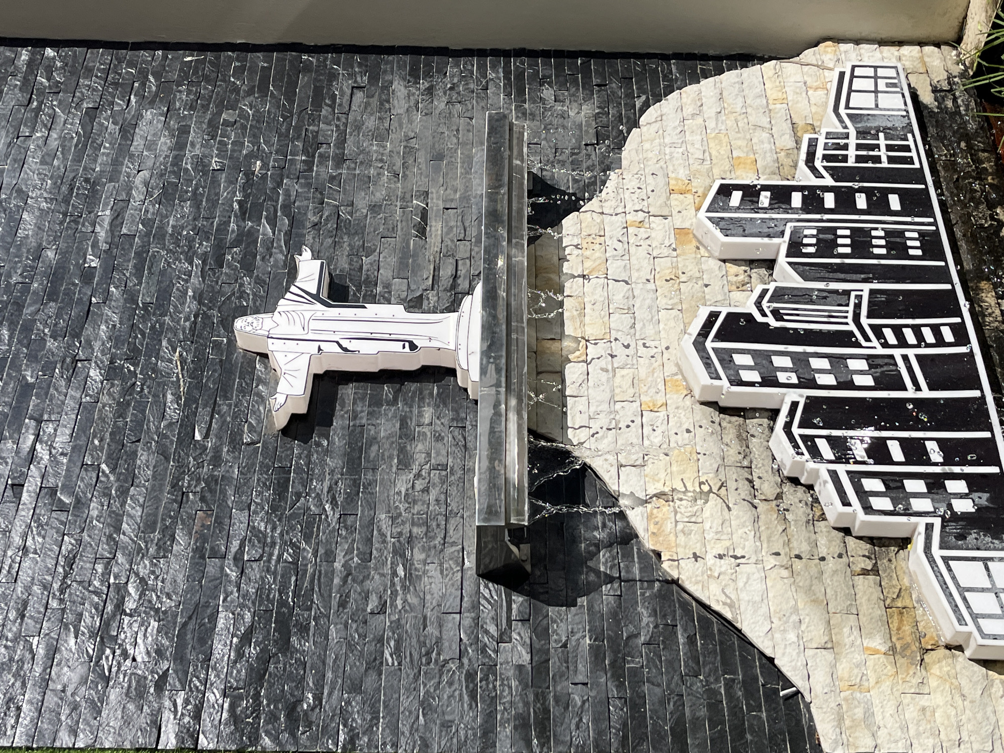 This image captures an outdoor architectural installation in an unidentified urban environment. The central visual element is a stone-tiled wall featuring a contrasting design. One side of the wall is covered in dark grey, horizontally arranged stone tiles, while the other section consists of lighter, beige-toned tiles. A metallic, rectangular structure bisects the wall vertically.
Attached to the dark grey section is a white, abstract sculpture resembling a futuristic aircraft. Adorning the lighter stone section is a black and white artwork depicting a stylized building or factory complex, composed of geometric shapes and patterns with distinct white borders. Water trickles down the metallic divider creating a cascade effect.
The environment suggests a modern outdoor setting, possibly part of a garden or decorative urban space. The composition highlights the textures of the stone and the interplay of colors and forms in the art installation.