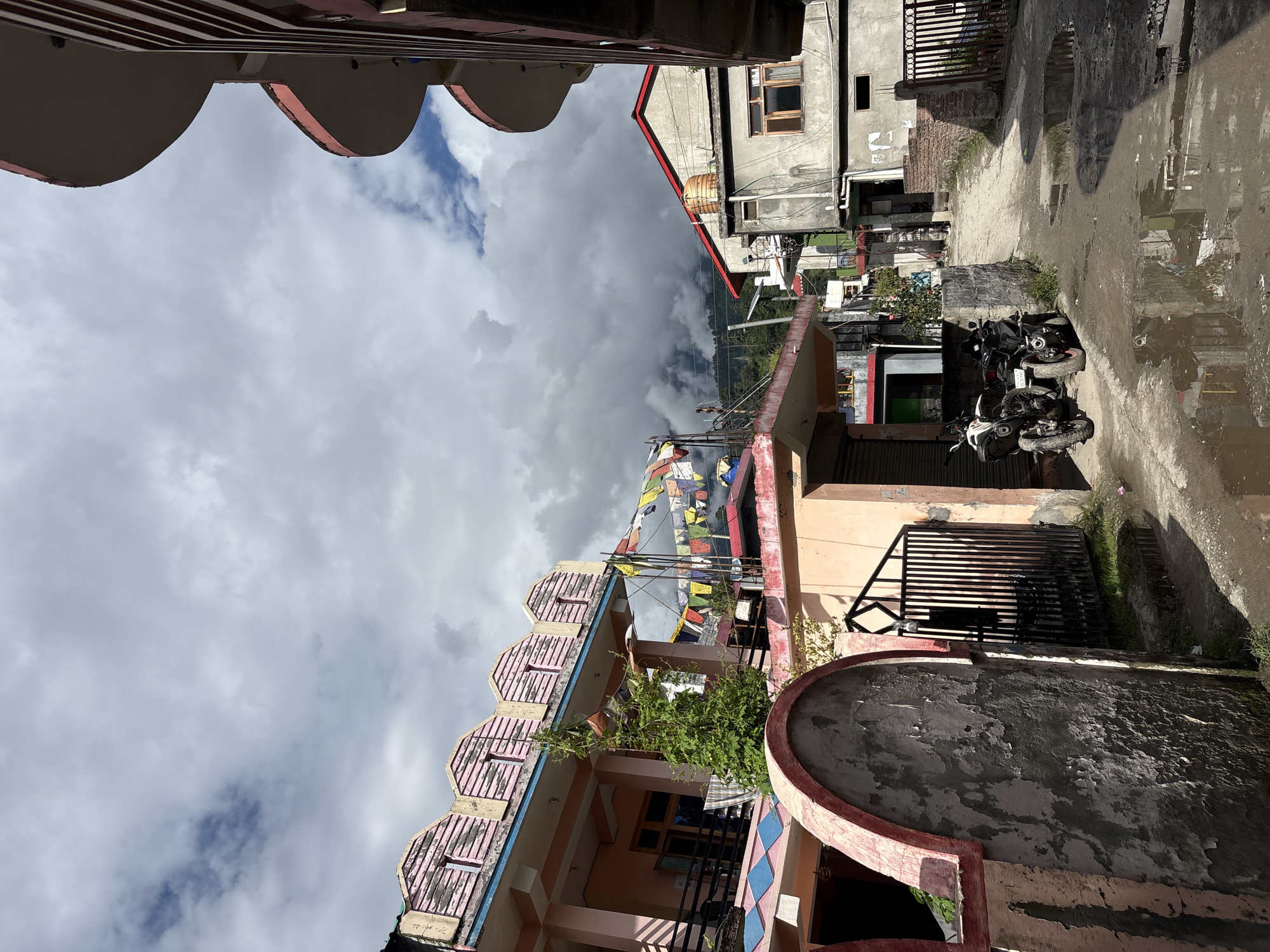 This is a street scene in Patlikuhal, India. The image depicts a narrow, unpaved street with buildings on either side. The sky is cloudy, with patches of blue visible. In the foreground, there are two motorcycles parked on the side of the road. The buildings are a mix of styles, with some featuring colorful decorations and others appearing more weathered. Prayer flags are strung across one of the buildings. The ground is wet, with puddles reflecting the surrounding structures. The overall impression is one of a vibrant, lived-in urban environment.
