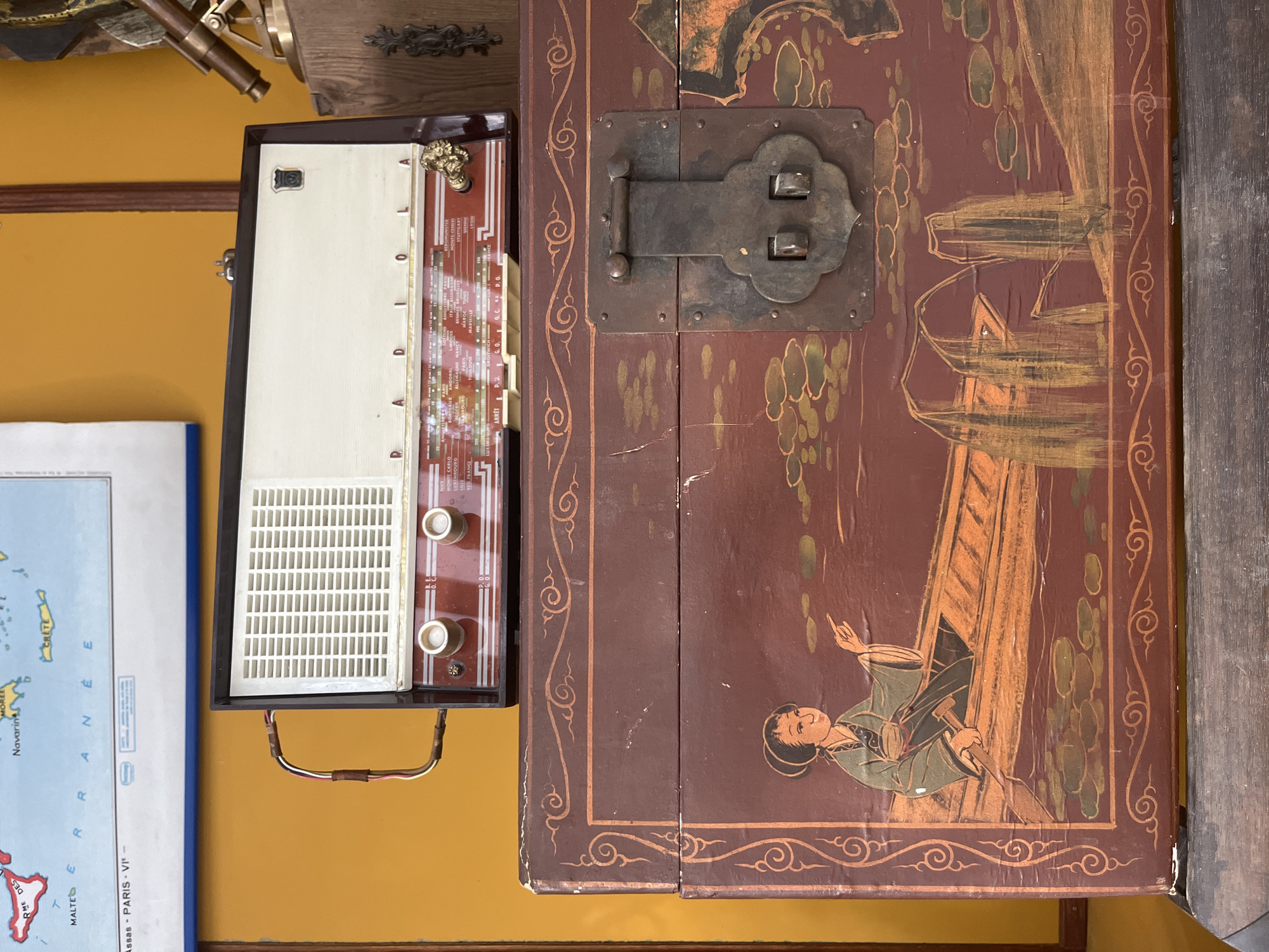 The image depicts a vintage radiola placed on top of a decorative wooden chest. The radiola is dark red and cream-colored with a white speaker grill and two prominent knobs on the front. A dial with various city names is visible beneath the speaker. The wooden chest is dark brown with intricate red and gold painted designs, including a depiction of an Asian figure in a boat on the front. A large, antique metal latch secures the chest. Behind the radiola, a portion of a map showing parts of Europe is visible against an orange wall. To the right of the radiola, a brass telescope on a wooden stand is partially visible. The overall scene suggests a vintage or antique collection displayed indoors. There is no discernible location in Geneva, Switzerland.