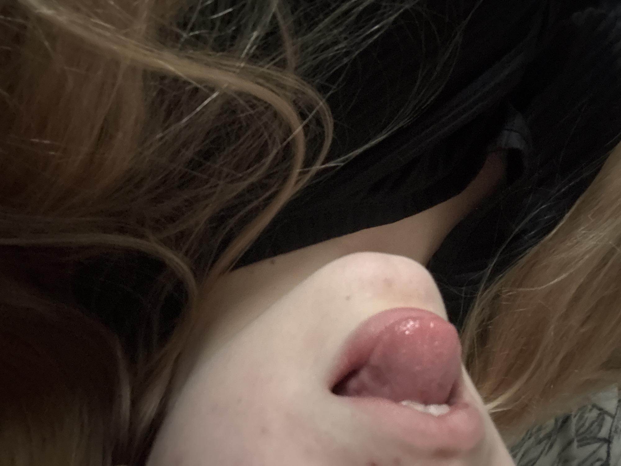 The media file is a close-up, low-angle, and upside-down shot of a person's mouth and chin, with their tongue sticking out. Their skin is pale with some light freckling visible. The person has blonde hair, which is partially visible framing the lower part of the image. They are wearing dark clothing, possibly a jacket or a shirt. The background is dark and out of focus, obscuring any details about the setting. The lighting appears to be dim, creating a moody atmosphere. There is no visible text in the image. The activity taking place is the person sticking out their tongue. There are no discernible cues about the time of day, weather, or specific location. The emotion conveyed is playful or suggestive.