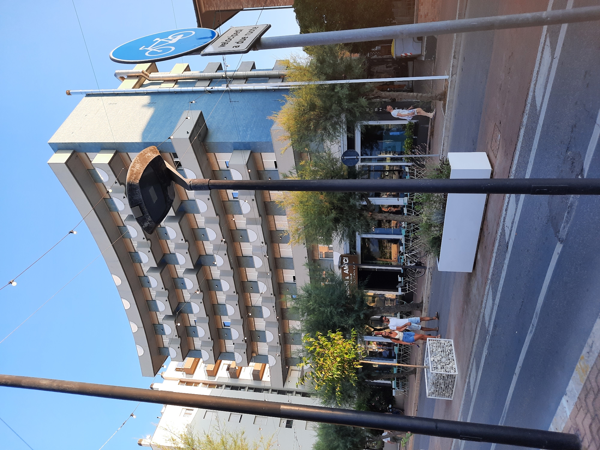 In Pesaro, Italy, a multistory building with a curved facade featuring multiple balconies is visible. A street lamp stands in the foreground, and a blue circular sign with a bicycle icon and accompanying text is mounted on a pole to the right. A sidewalk runs in front of the building, lined with trees and outdoor seating for a restaurant. A couple walks on the sidewalk, and other individuals are present. The scene suggests a street in a coastal or resort area, bathed in the warm light of late afternoon or early evening.